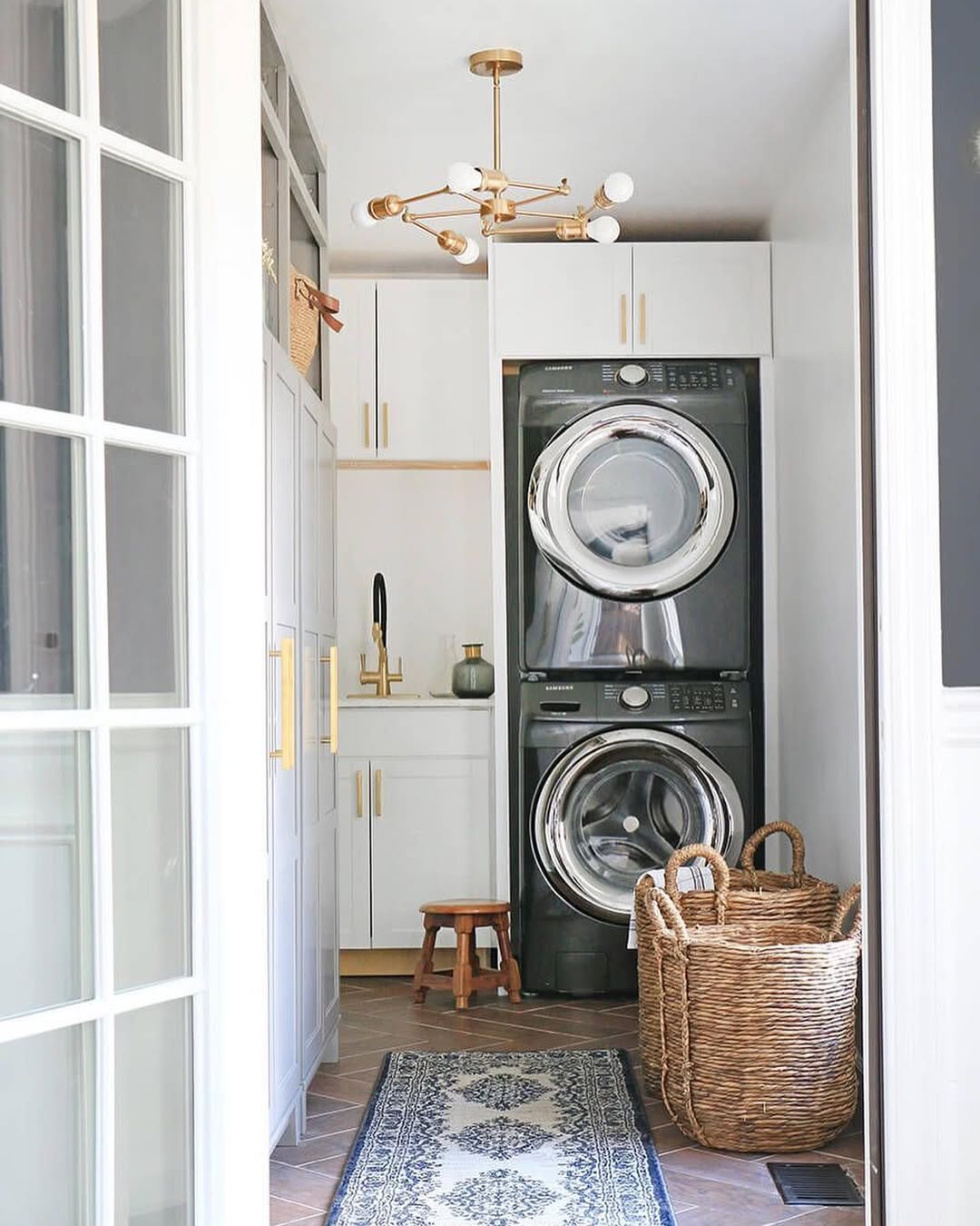 A modern Boho laundry room features stacked washer and dryer, white cabinets, a rug, a wicker basket, a stool, and a gold light fixture—perfect for anyone seeking unique laundry room ideas with boho decor charm.