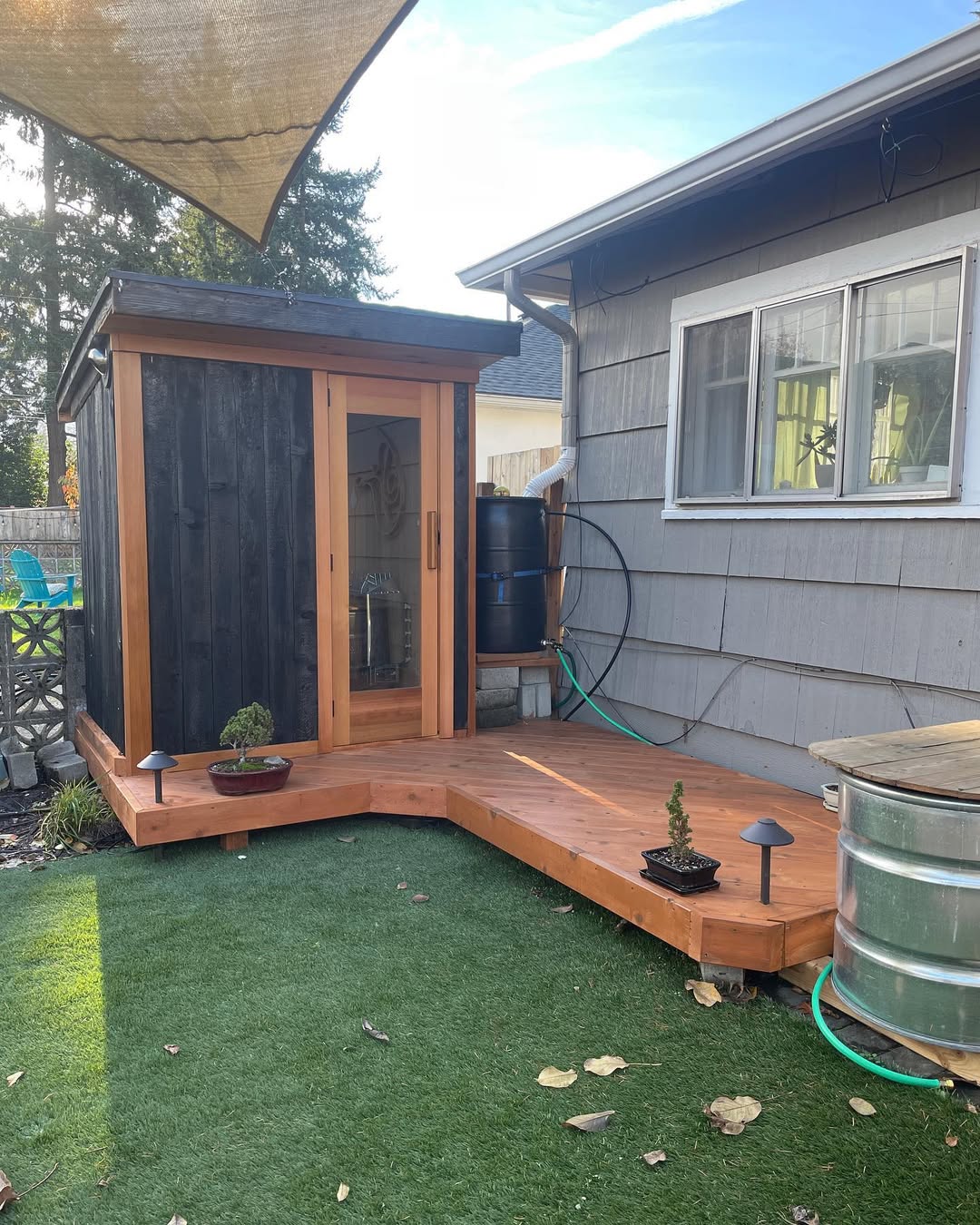 A small wooden backyard sauna with a glass door sits on a raised deck beside a gray house, surrounded by artificial grass and garden decorations—perfect inspiration for outdoor sauna ideas.
