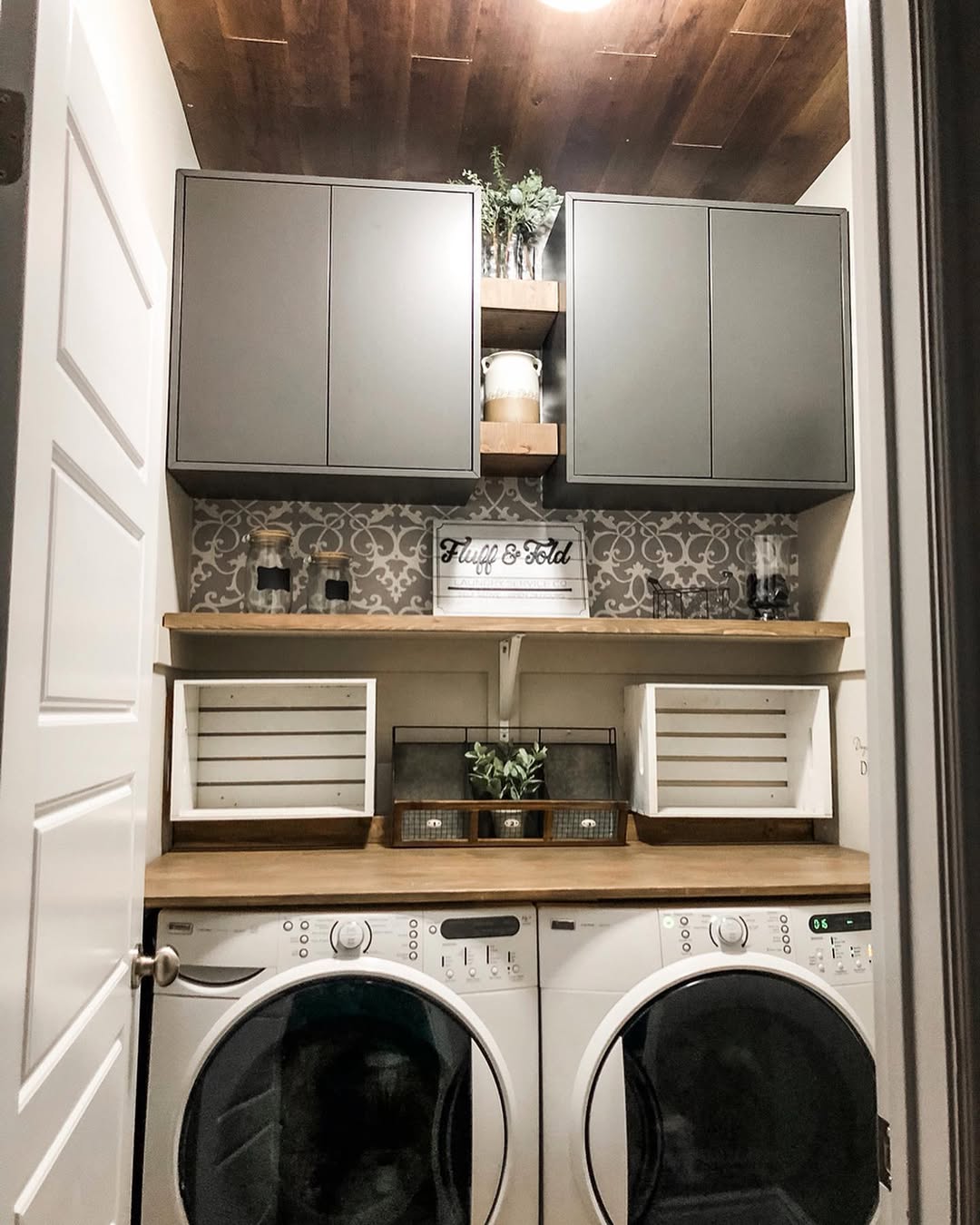 A compact laundry room with a front-loading washer and dryer, wooden shelves, gray cabinets, and decorative items neatly arranged above—perfect inspiration for small laundry spaces.