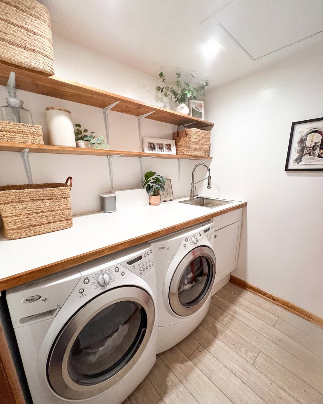 A cozy small laundry room features a white washer and dryer, wooden shelves with baskets, and a countertop with a sink. Decor is complemented by framed photos and plants, making efficient use of every inch.