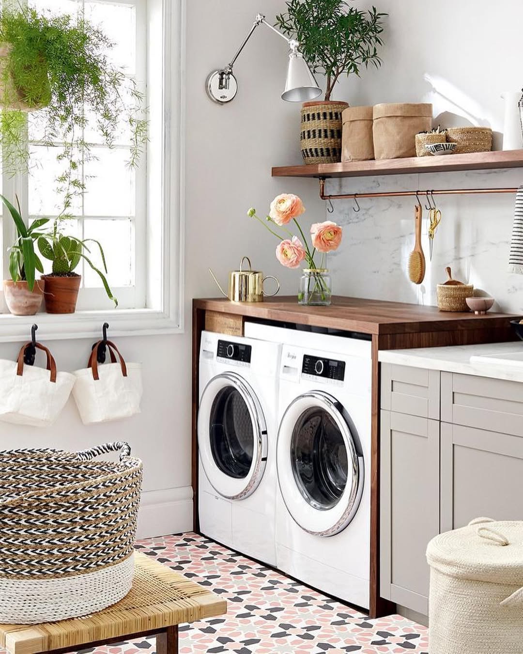 A bright, modern Boho laundry room with a washer and dryer, wood countertop, potted plants, woven baskets, and a pink patterned tile floor—perfect inspiration for fresh laundry room ideas.