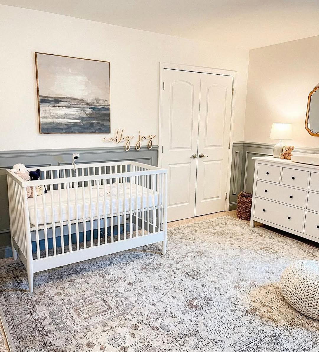 A tidy nursery with a white crib, stuffed animals, dresser, lamp, and framed wall art, featuring light gray paneling and a patterned rug—perfect for first-time moms seeking baby boy nursery ideas in 2025.