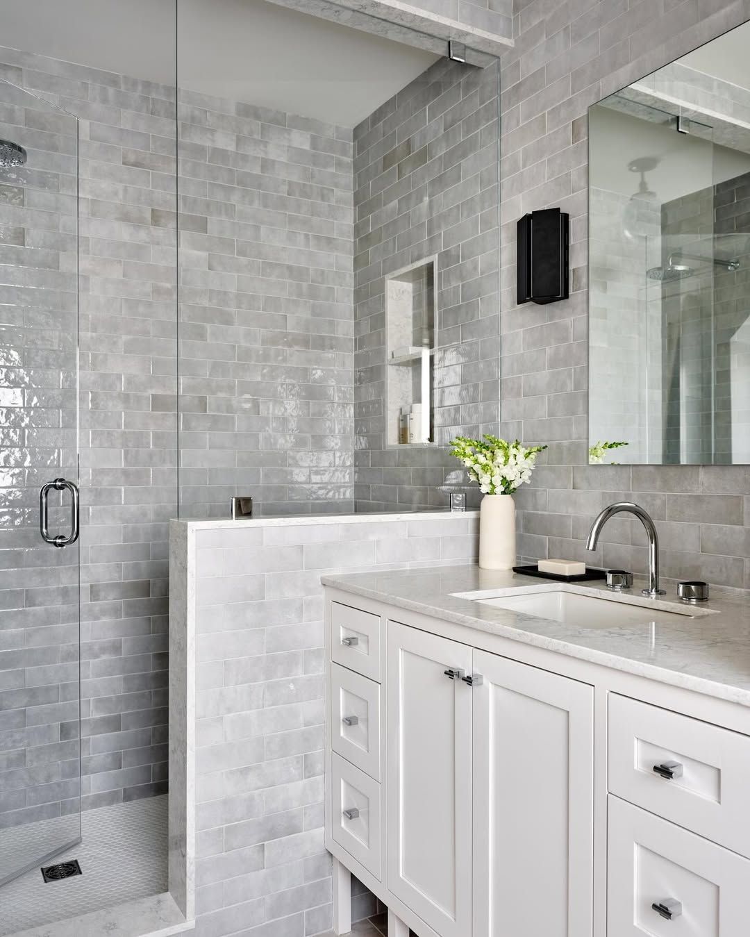 Modern bathroom with gray tiled walls showcasing stylish bathroom tile ideas, a glass-enclosed shower, white vanity with drawers, rectangular mirror, and a vase with white flowers on the countertop.