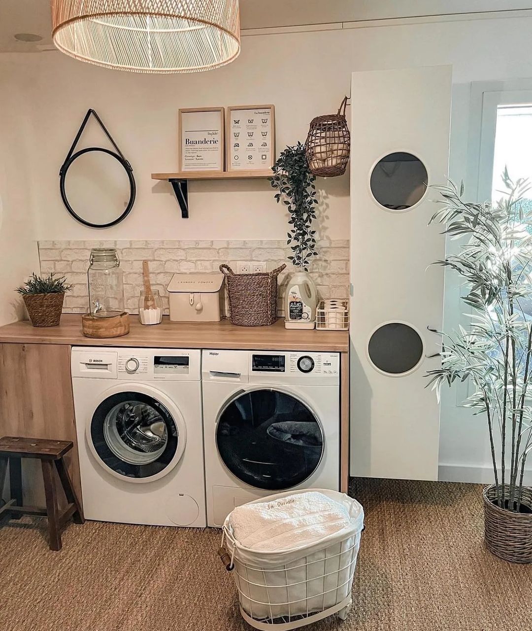 Modern boho laundry room with a washing machine and dryer, countertop, wicker baskets, wall decor, a mirror, plants, and a white laundry hamper on a woven floor—perfect for those seeking stylish laundry room ideas.