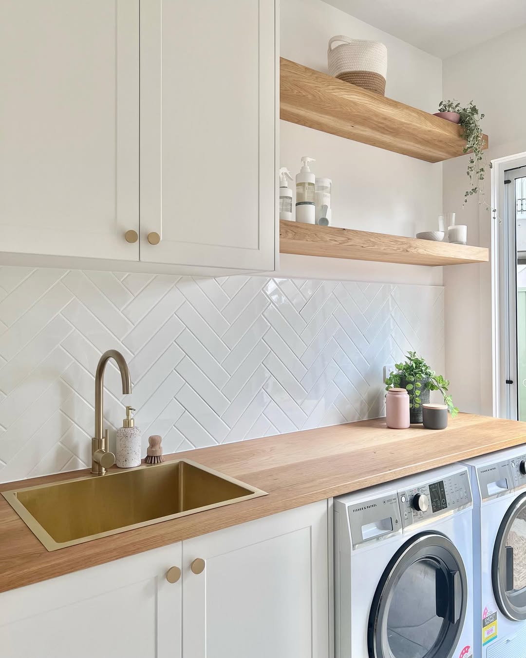 Modern boho laundry room with a wood countertop, brass sink, white cabinets, open shelves, and side-by-side washer and dryer. White herringbone tile backsplash and plants complete the bohemian laundry room vibe.