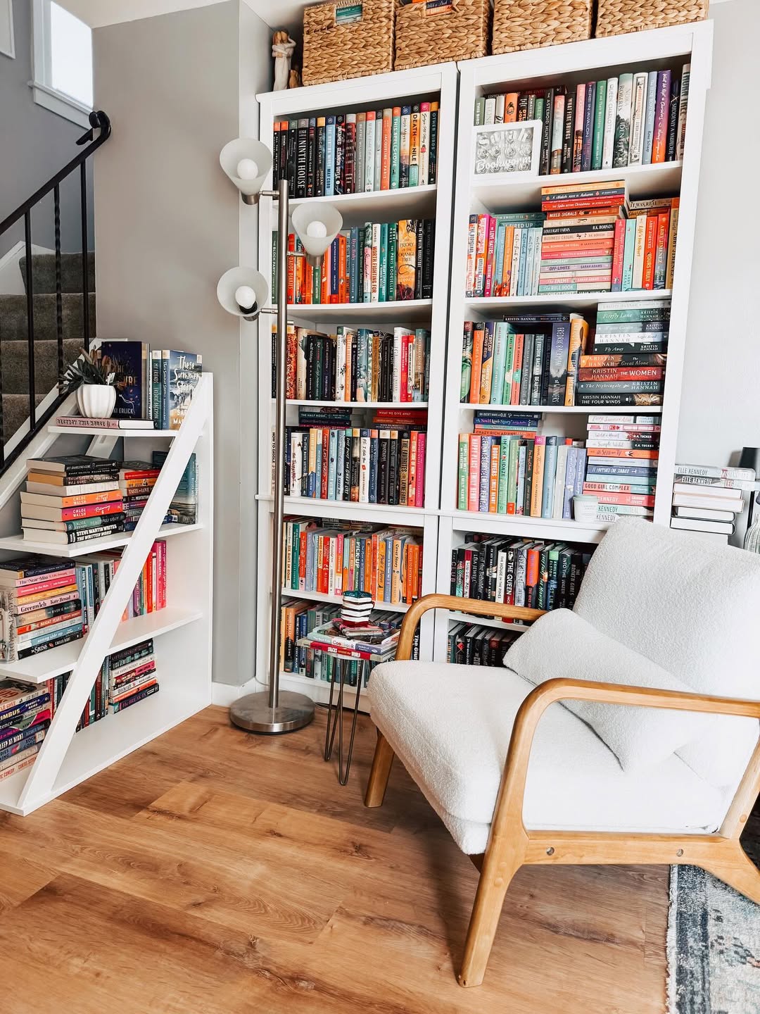 A cozy reading nook with a white armchair, side table stacked with books, two tall white bookshelves filled with books, and a small bookshelf under the staircase—perfect for book nook ideas.