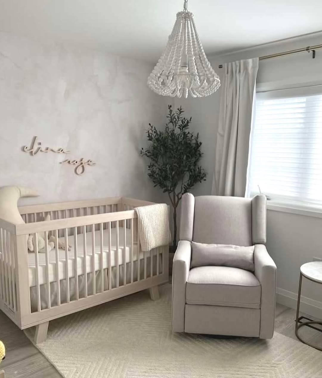 A neutral-toned nursery with a light wooden crib, a beige armchair, a faux plant, and a beaded chandelier beside a window with white curtains—perfect for first-time moms seeking chic nursery ideas.