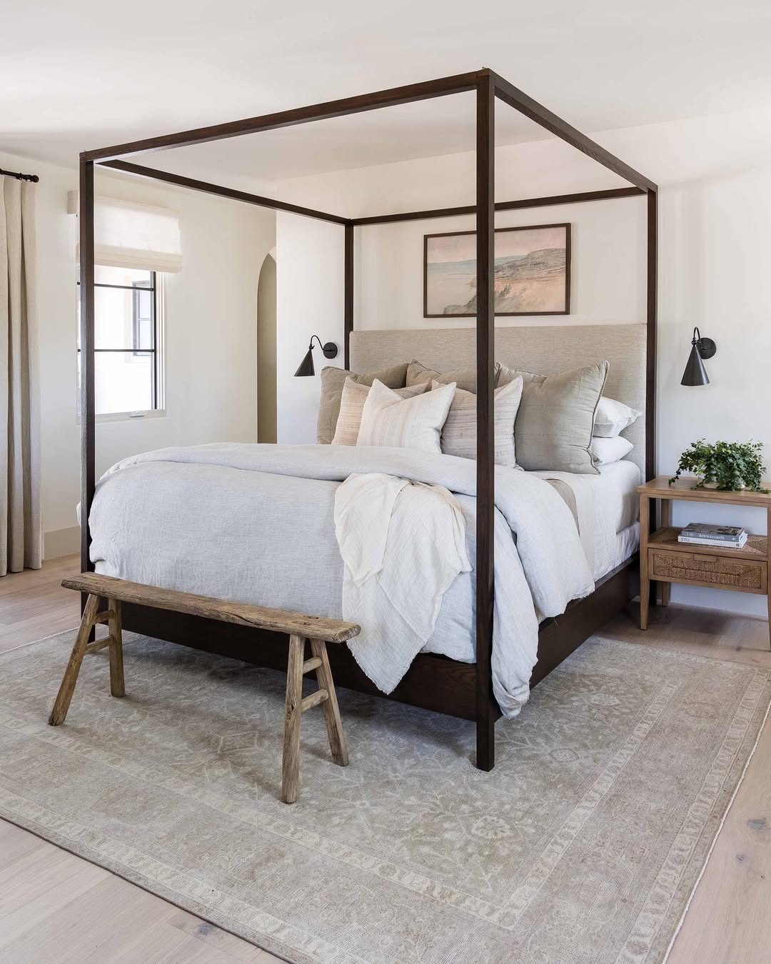A modern bedroom perfect for Interior Lovers, featuring a four-poster bed, neutral bedding, a wooden bench at the foot, a nightstand with a plant, and a wall-mounted lamp—an inspiring space for 2025 Bedroom Color Schemes on a large patterned rug.