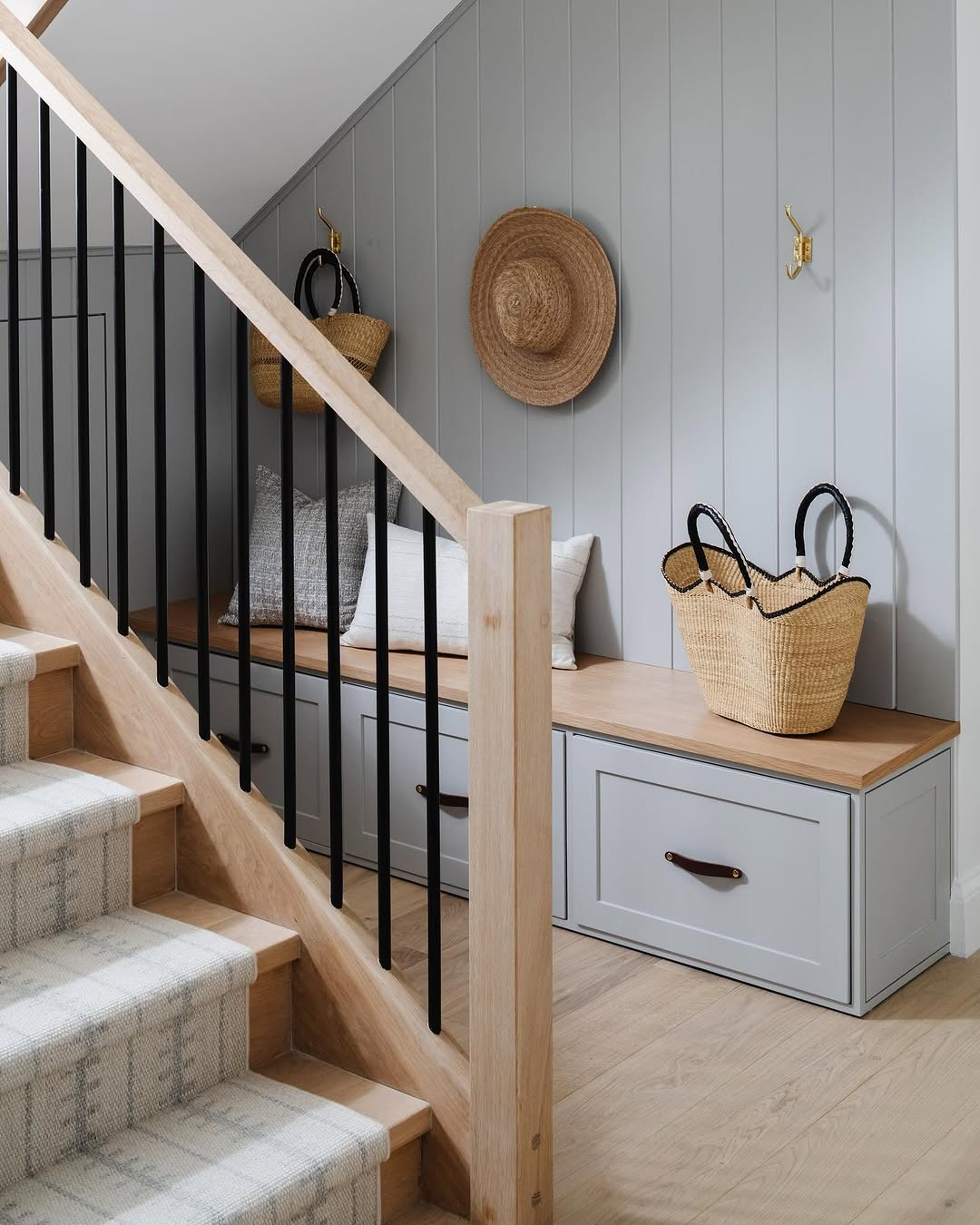 A wooden staircase with a carpet runner leads to a cozy nook featuring clever under stairs storage—built-in bench, gray drawers, cushions, straw bags, and a straw hat hanging on a light gray paneled wall.