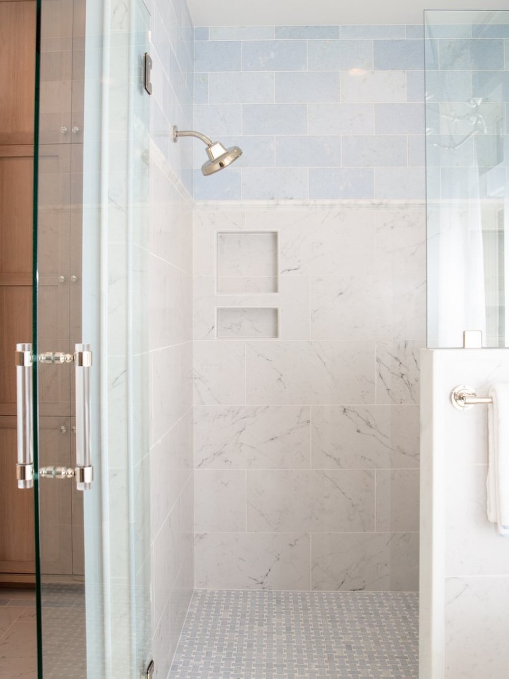 Glass door shower with marble-look tile walls, built-in shelves, silver showerhead, and small light blue floor tiles. For spa-worthy bathroom decor, a white towel hangs on the adjacent wall. Great inspiration for bathroom tile ideas.