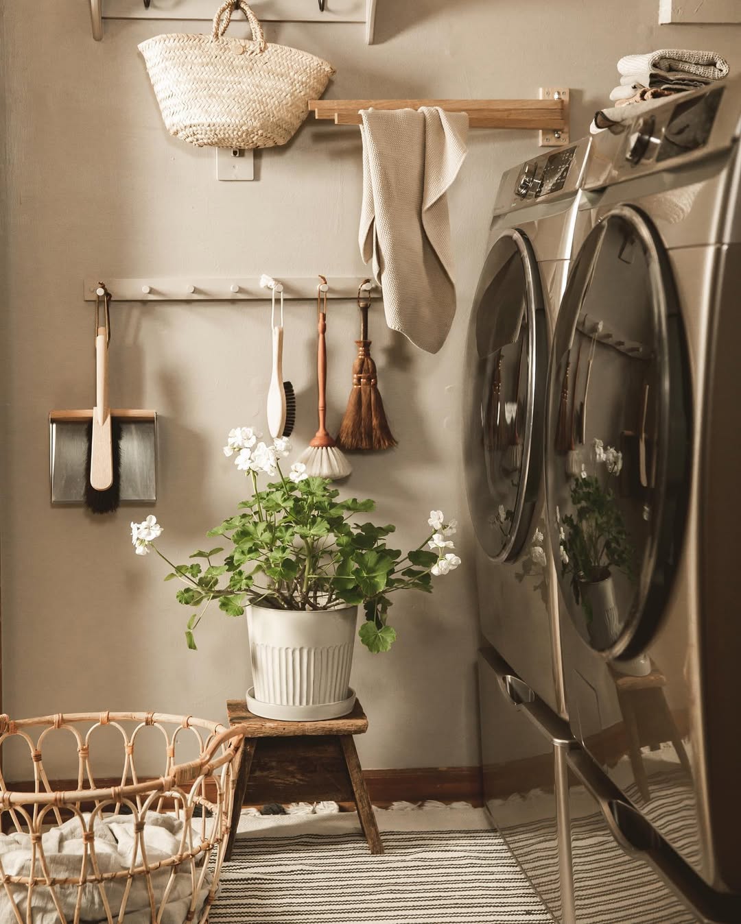 Small laundry room featuring a washer and dryer, broom and brushes on wall hooks, a potted plant on a stool, a woven basket, and a striped rug—perfect for inspiring laundry room organization.