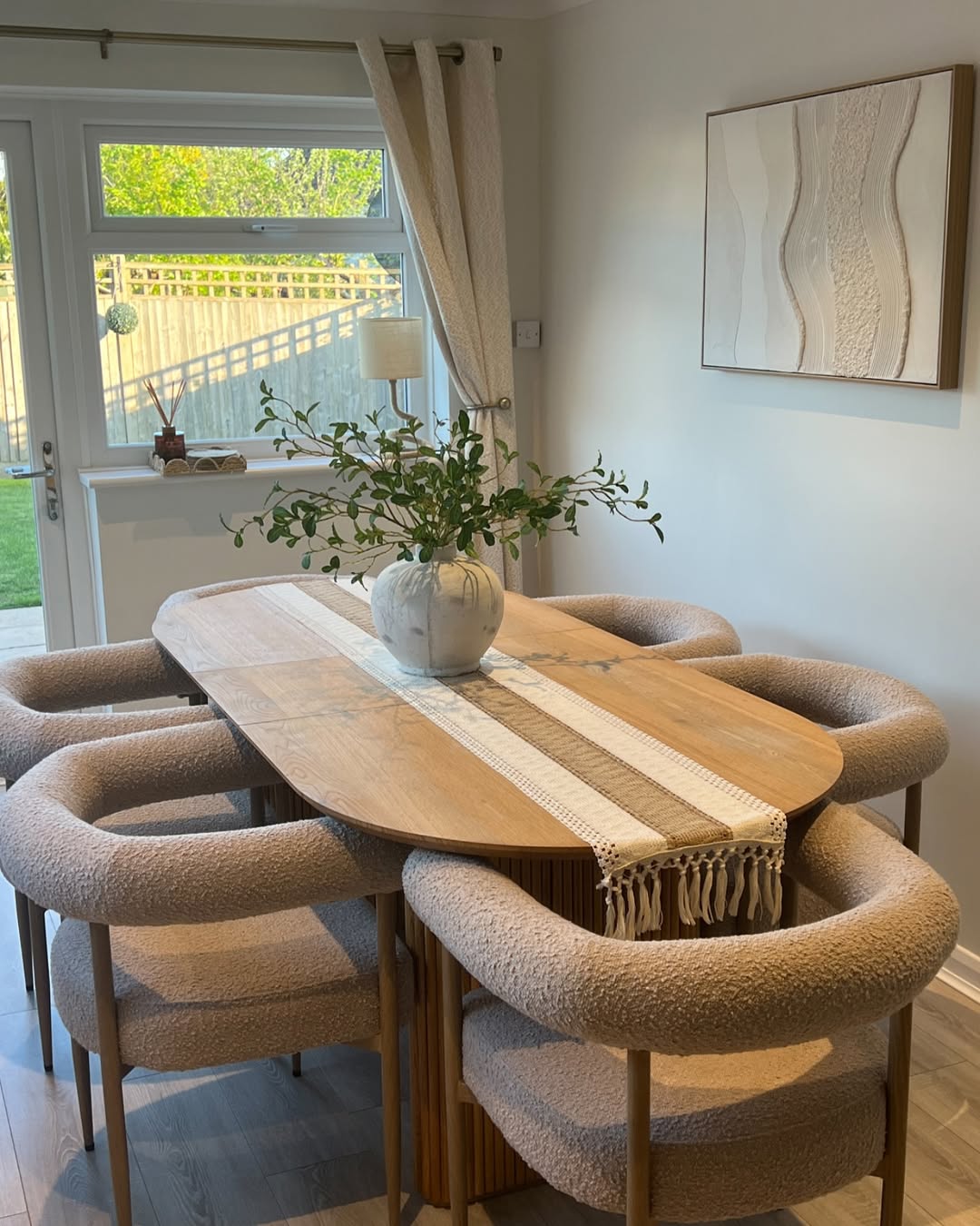 A wooden dining table with six cushioned chairs, a woven table runner, and a white vase featuring stylish vase decor as the centerpiece, in a bright room with large windows and neutral decor.