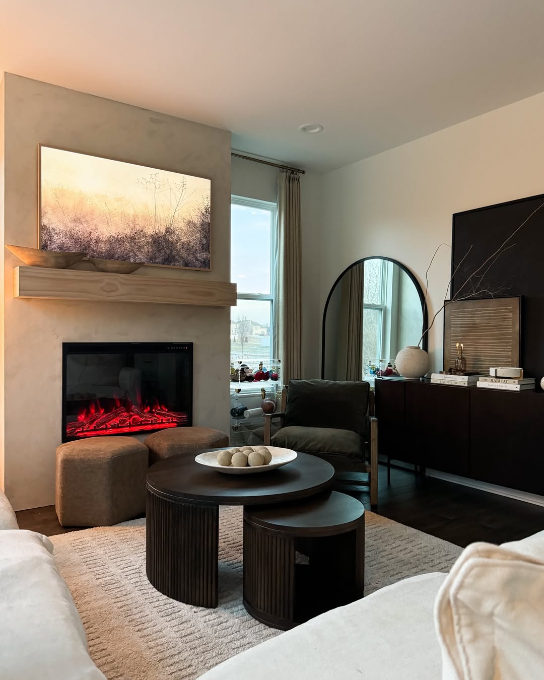 A modern living room featuring organic modern touches with a wall-mounted TV above an electric fireplace, round nested coffee tables, a large round mirror, and neutral-toned decor—perfect for inspiring fresh living room ideas.