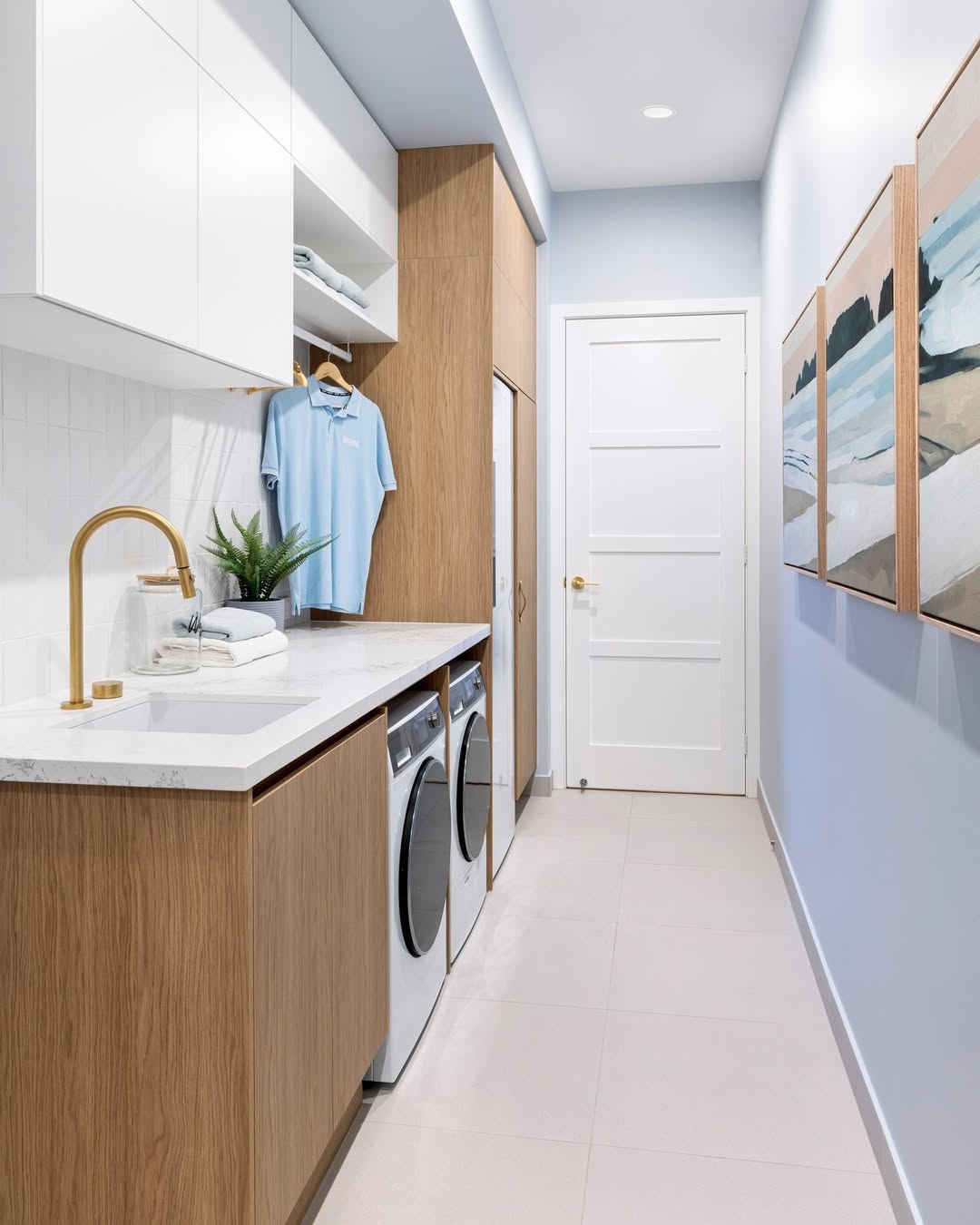 Modern stacked laundry room with washer and dryer, wooden cabinets, white countertops, a sink with a gold faucet, hanging shirt, towels, and wall art on a light blue wall.