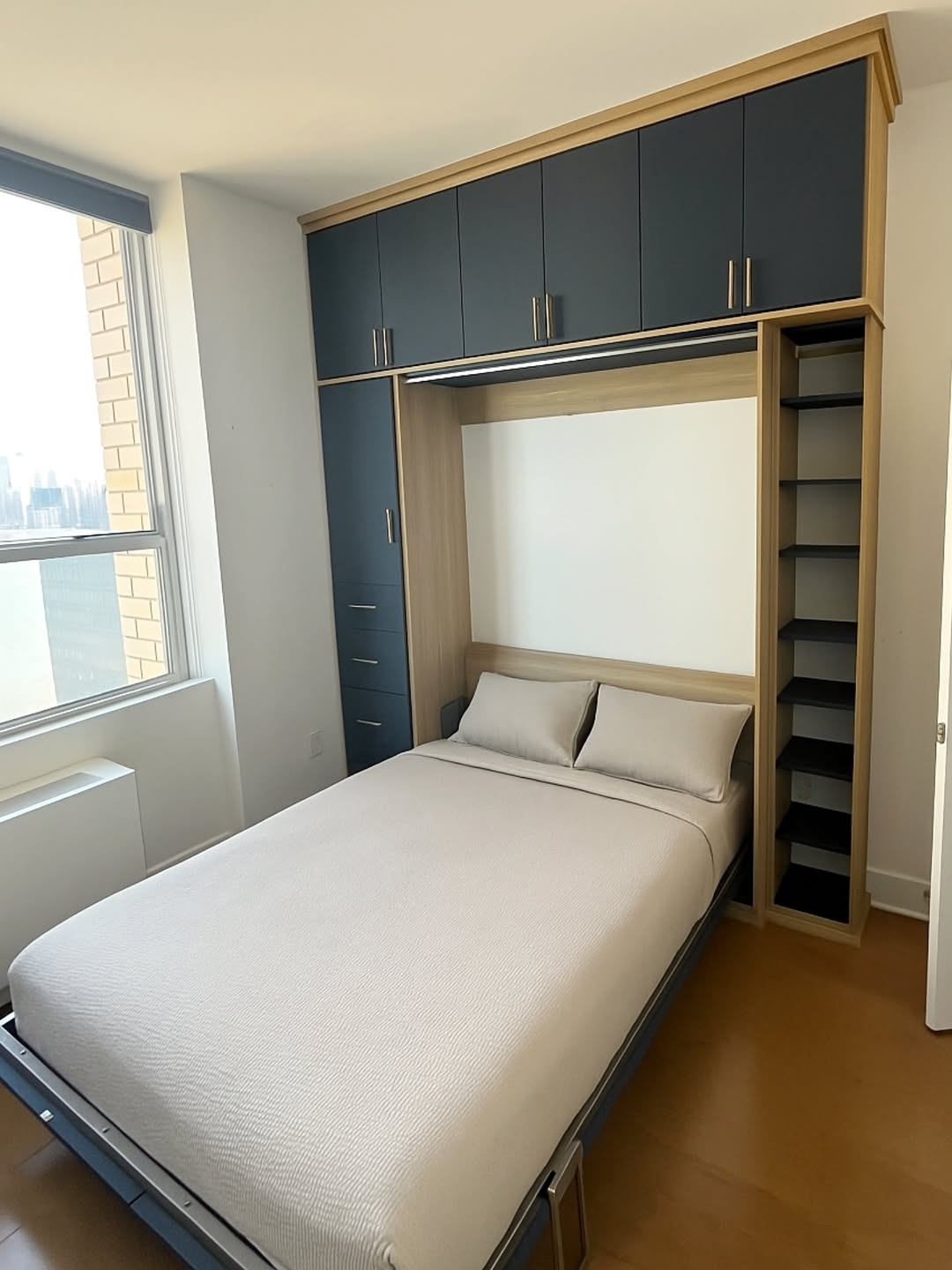 Queen-sized bed in a modern room with built-in overhead cabinets, open shelving, and drawers; large window with a city view on the left—perfect for those seeking stylish Murphy bed ideas and smart bed design solutions.