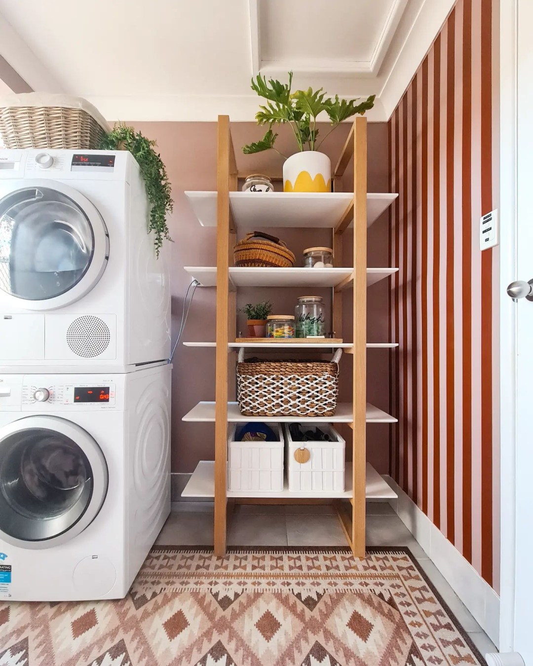A boho laundry room featuring a stacked washer and dryer beside a wooden shelving unit with baskets, boxes, and plants. A zigzag patterned rug completes the cozy space—perfect inspiration for laundry room ideas.