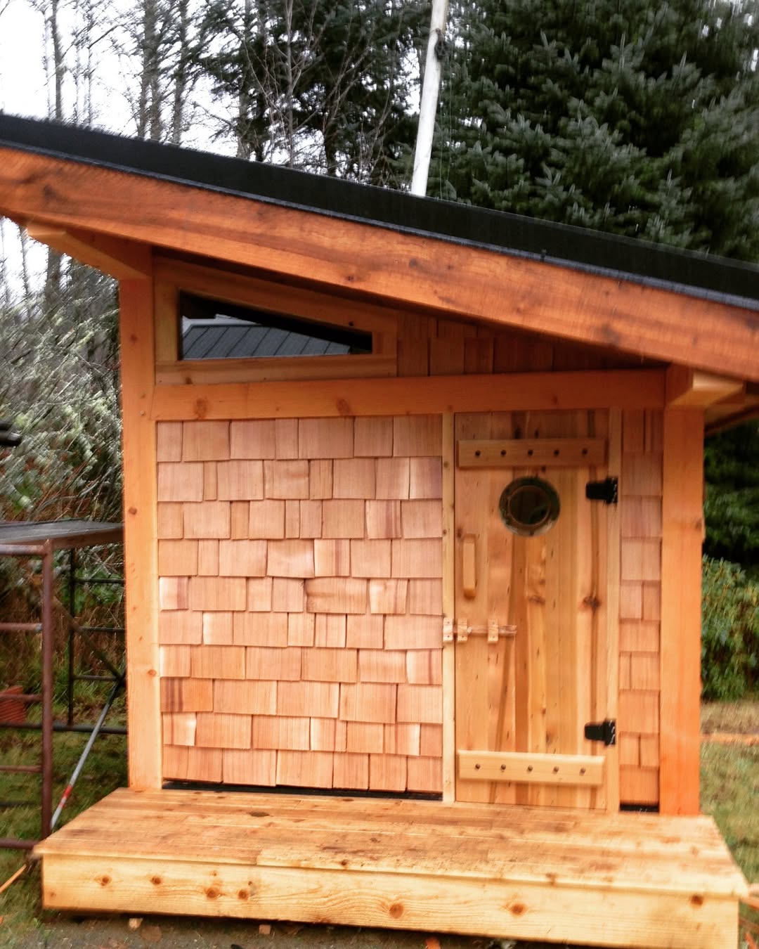 A small wooden shed with cedar shingles, a slanted roof, and a door featuring a round window and black hinges, perfect as a backyard sauna and offering inspiration for gorgeous outdoor sauna ideas, set outdoors near trees.