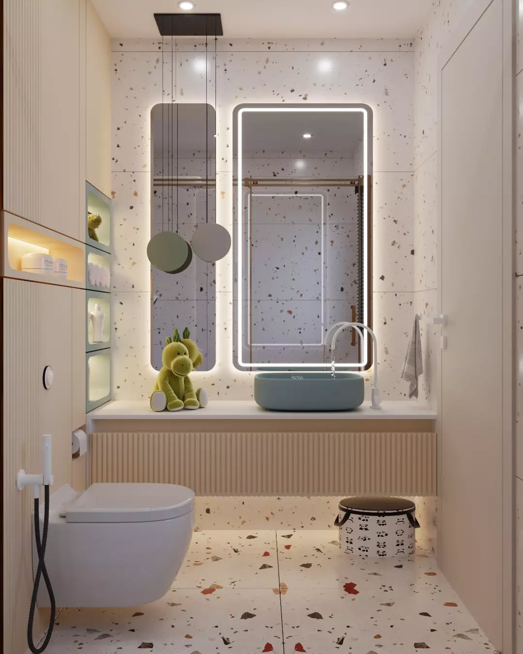 Modern kids bathroom with terrazzo walls and floor, wall-mounted toilet, illuminated rectangular mirror, blue sink, green plush toy on the counter, and built-in shelving with toiletries.