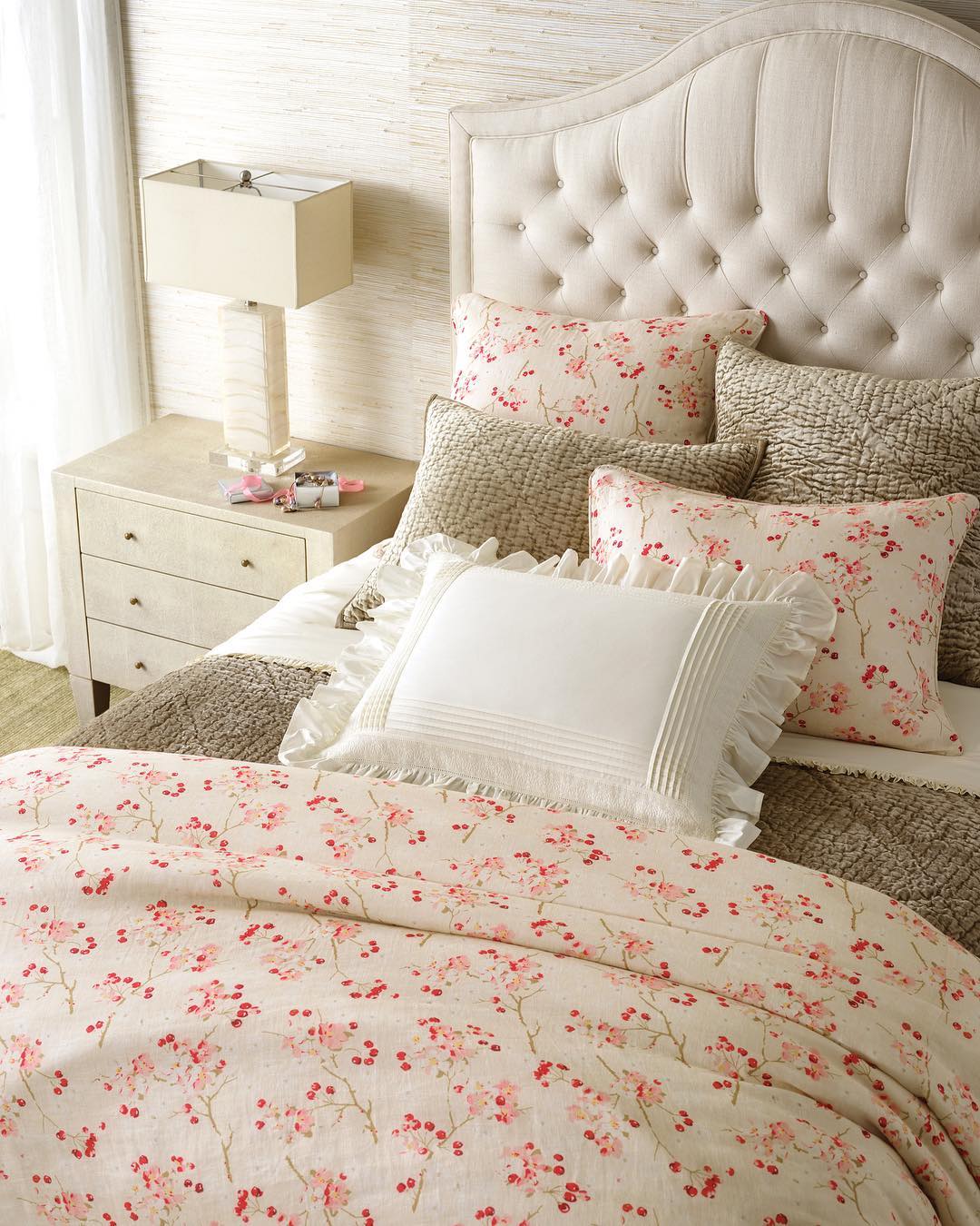 A neatly made bed with floral bedding and ruffled pillows stands beside a cherry wood bedroom table holding a lamp, clock, and books in a softly lit bedroom.