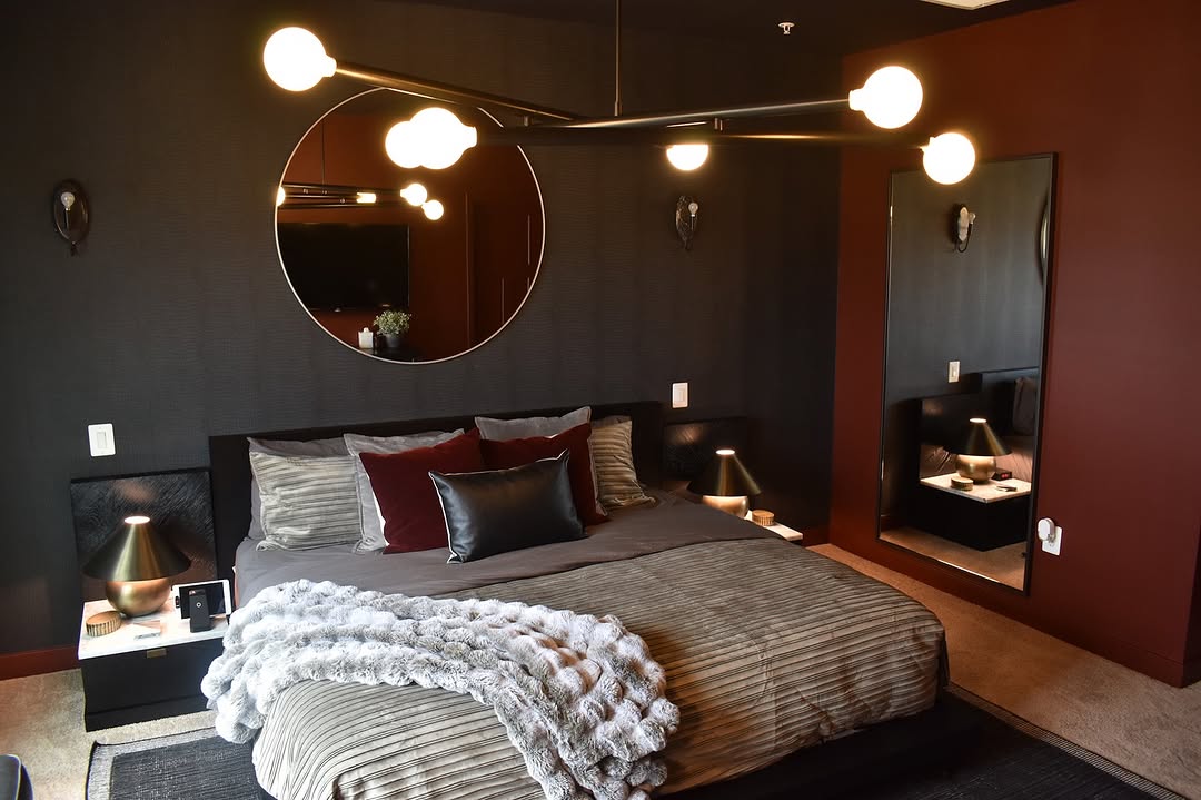 Modern dark feminine bedroom featuring a large round mirror above the bed, a geometric light fixture, deep-hued walls, two nightstands, and a textured throw blanket for added elegance.