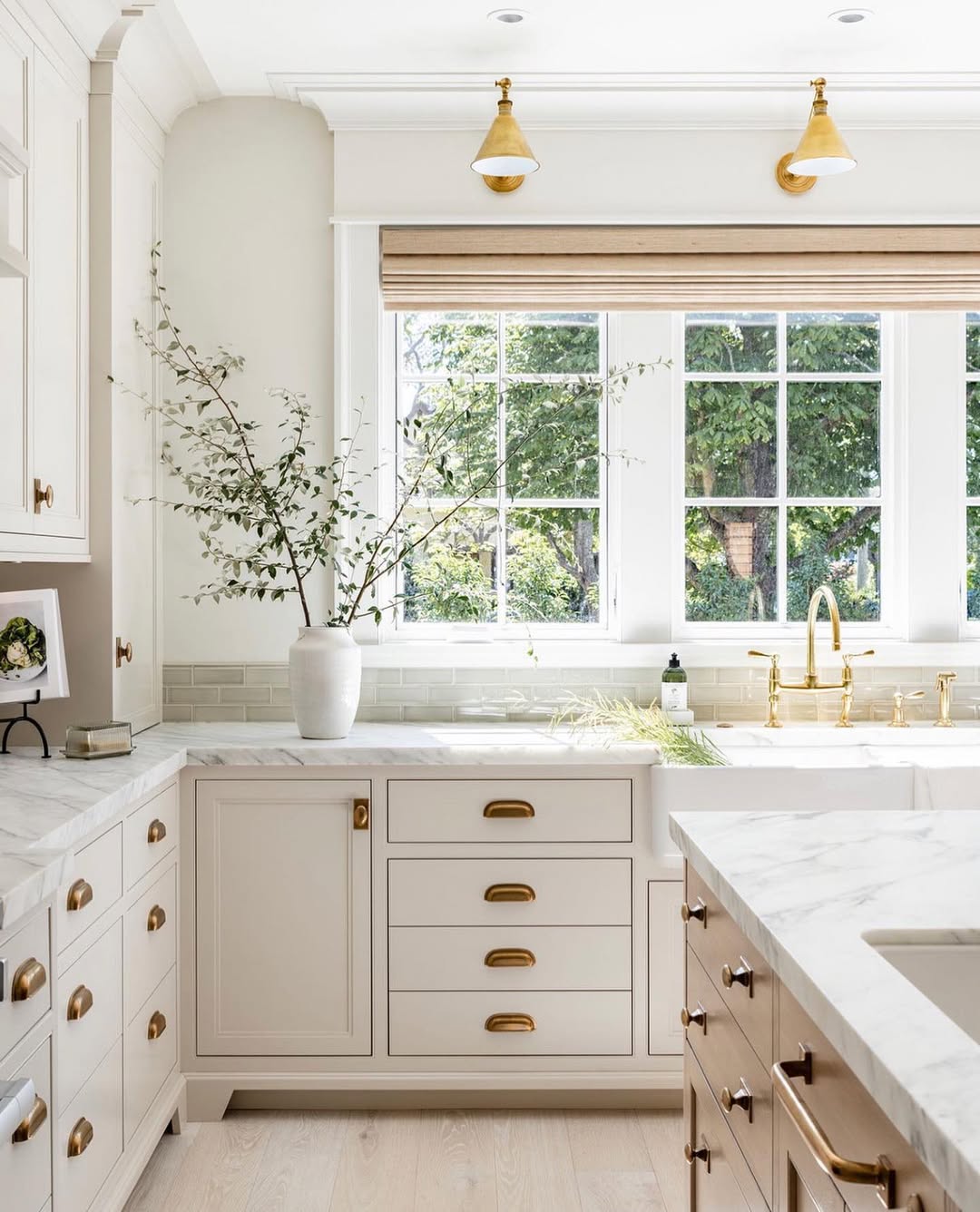 Bright, modern kitchen with white cabinets, marble countertops, brass hardware and fixtures—a showcase of stunning kitchen colors and fresh kitchen color ideas for 2025—plus a large window and a vase with branches on the counter.