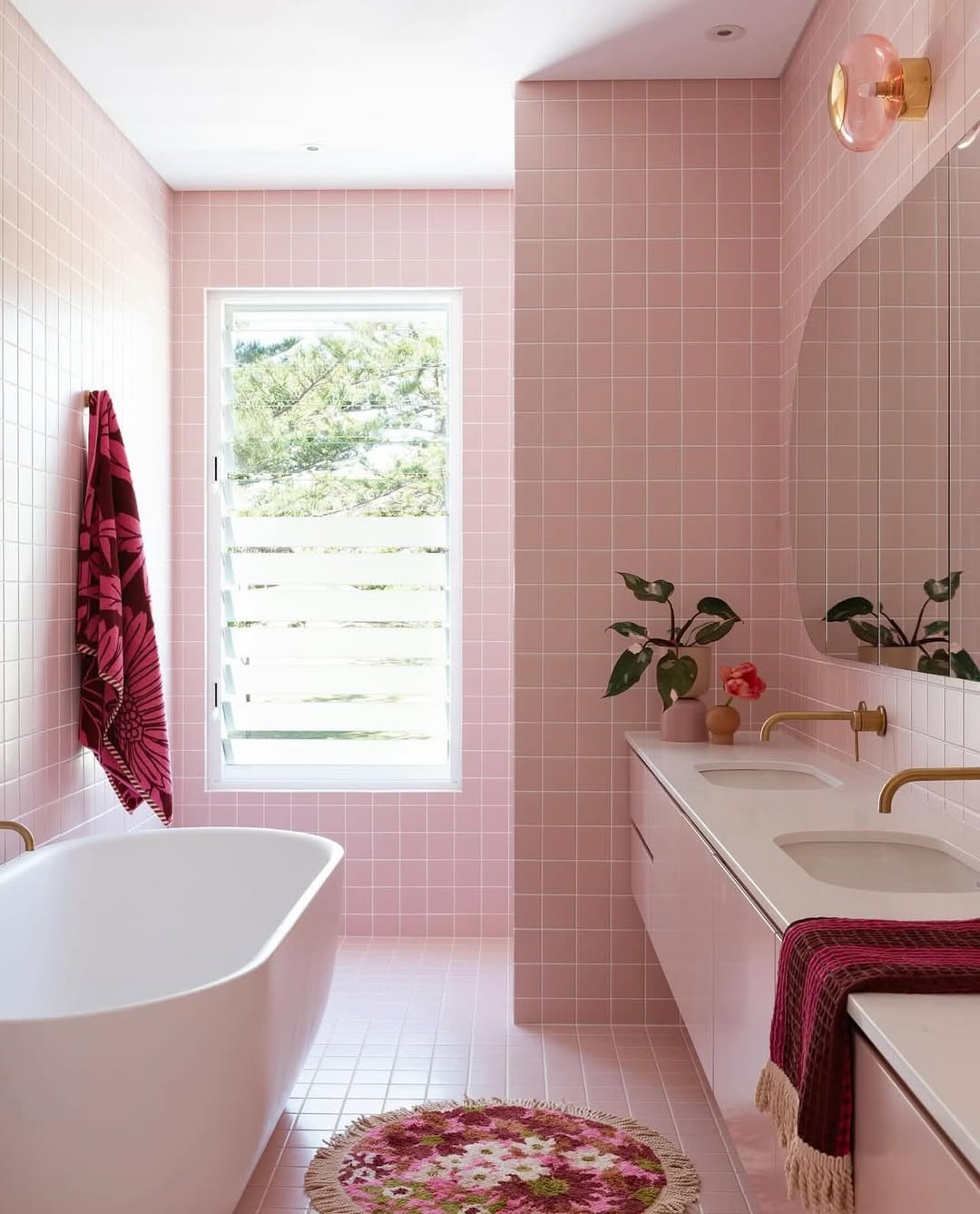 A pink bathroom with tiled walls and floor features a white bathtub, double sink vanity, gold fixtures, a potted plant, and pink decorative towels and rug. Natural light streams in through the window.