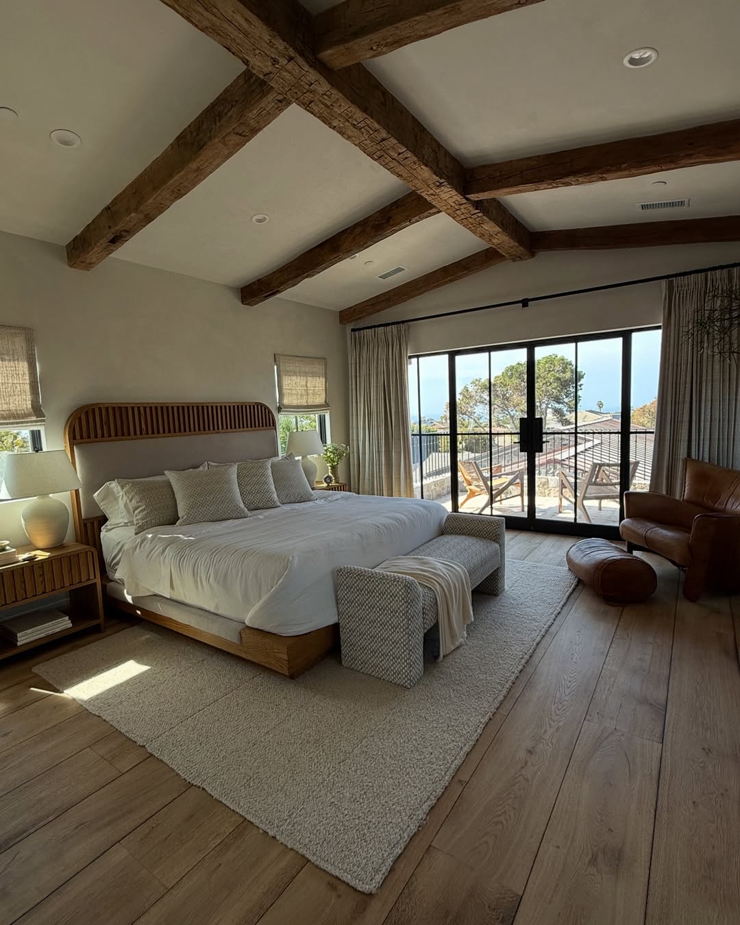 Spacious rustic bedroom with exposed wooden beams, a large bed, armchair, and glass doors opening to a balcony with outdoor seating and a view of trees—perfect for those seeking rustic bedroom ideas.