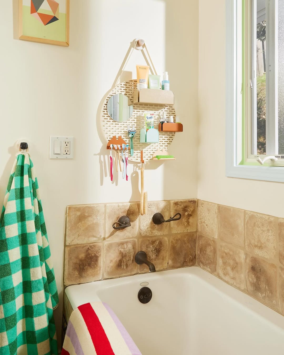 A kids bathroom featuring a bathtub with a striped towel, beige tiled walls, a green and white towel on a hook, and a hanging organizer with toiletries and toothbrushes below a window.