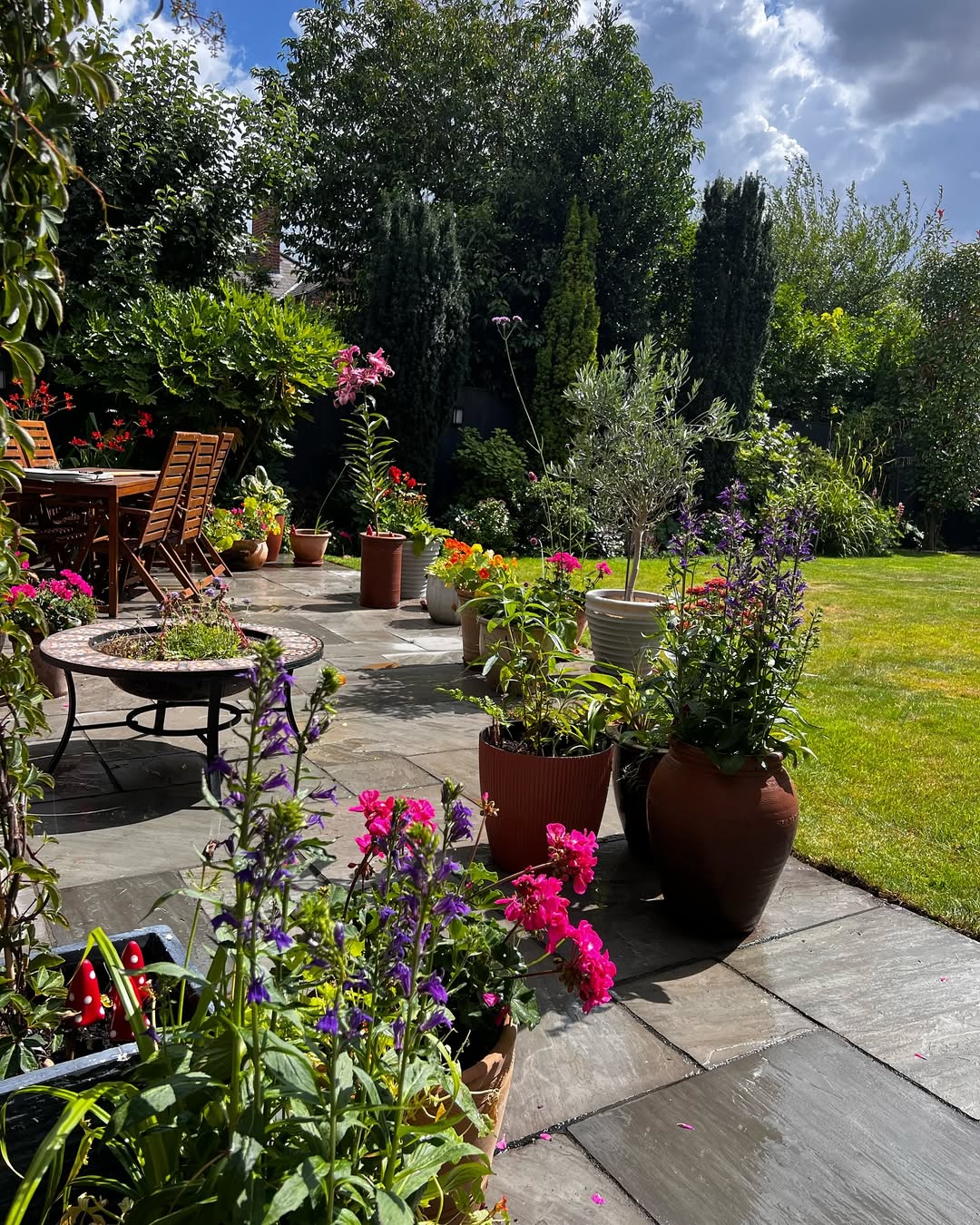A garden with colorful flowers in pots, wooden chairs and table on a stone patio, and a green lawn bordered by trees under a partly cloudy sky—perfect inspiration for Garden Ideas 2025.