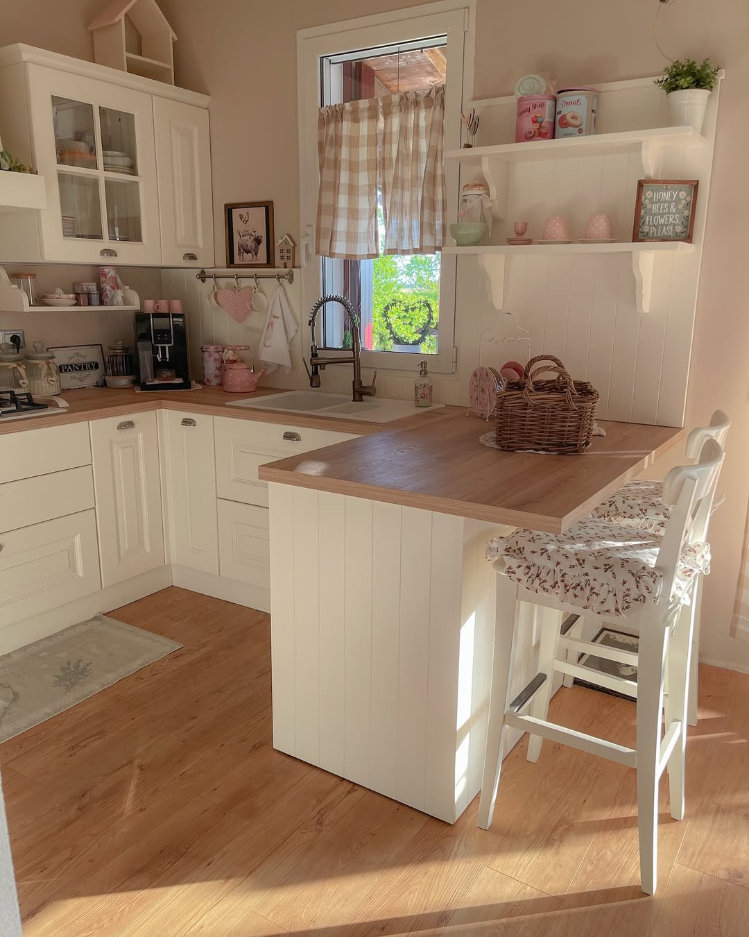 A cozy, sunlit farmhouse kitchen with white cabinets, wooden countertops, a coffee machine, wicker basket, and two cushioned chairs by a breakfast bar. Checkered curtains hang at the window—perfect inspiration for charming kitchen ideas.