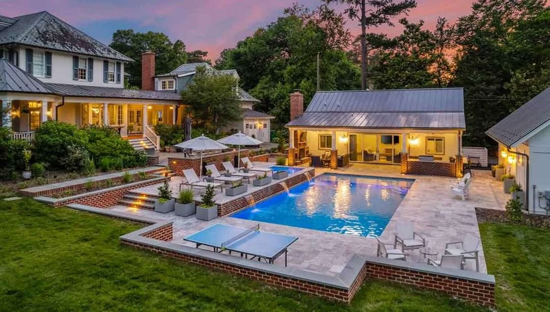 A large backyard with a rectangular swimming pool, lounge chairs, umbrellas, seating areas, and three houses surrounded by trees at sunset—perfect for anyone seeking backyard pool ideas.