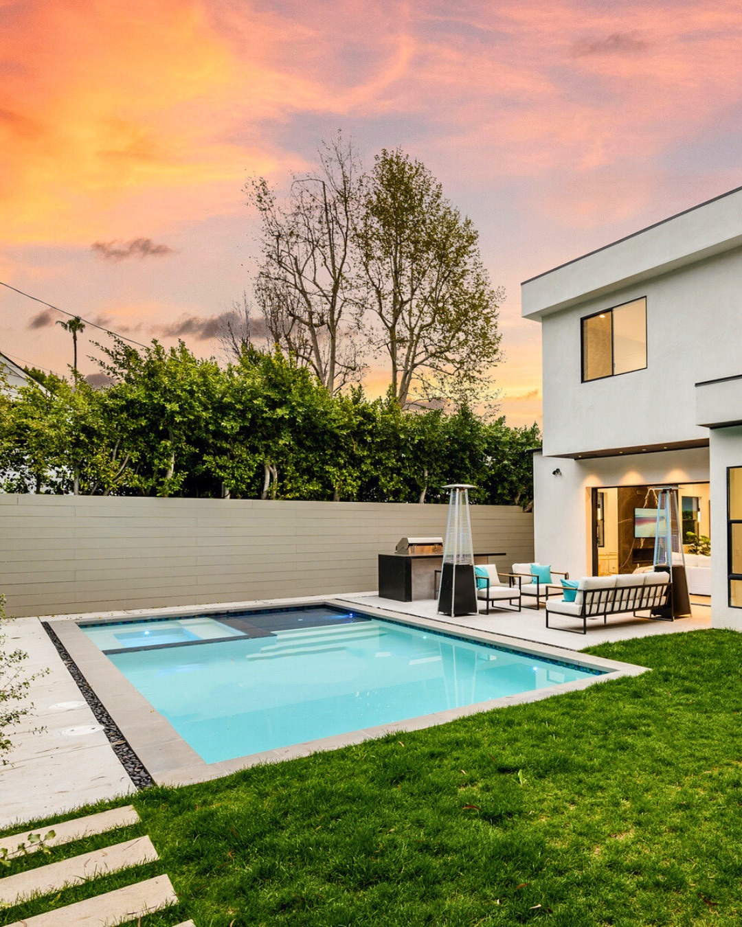 Modern backyard with a rectangular swimming pool, seating area, and green lawn—perfect for those seeking cool pool ideas and inspiration for stylish backyard pools—next to a white two-story house at sunset.