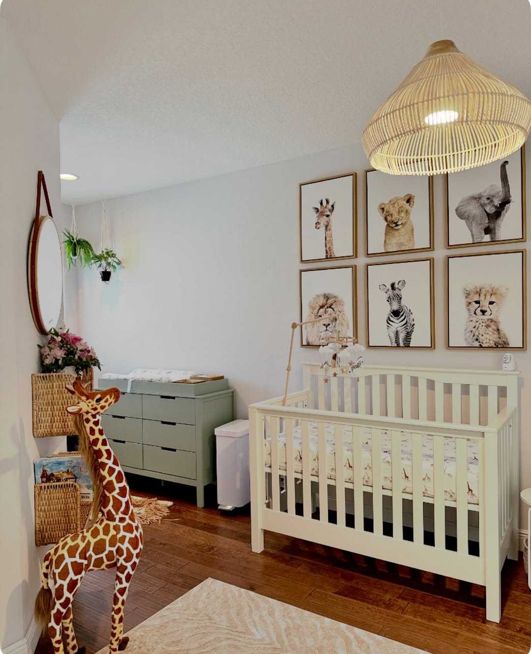 A perfect 2025 baby boy nursery idea for a first-time mom: a white crib, animal-themed wall art, a large giraffe plush, wicker baskets, a round mirror, and a green dresser under hanging plants.
