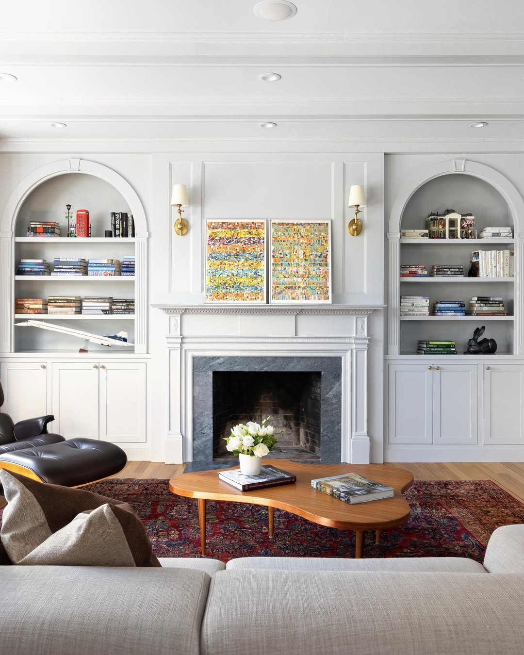A modern living room with fireplace built-ins, white shelves, a cozy fireplace, colorful art above the mantel, a wooden coffee table, and a patterned red rug.