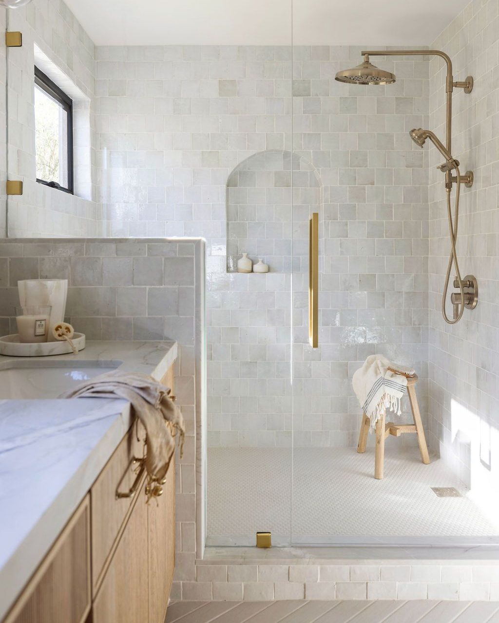 Modern bathroom with a glass-enclosed shower, rainfall showerhead, built-in niche, wooden stool with a towel, marble countertop, and light-colored tiles—perfect for creating a spa-worthy escape and inspiring new bathroom tile ideas.