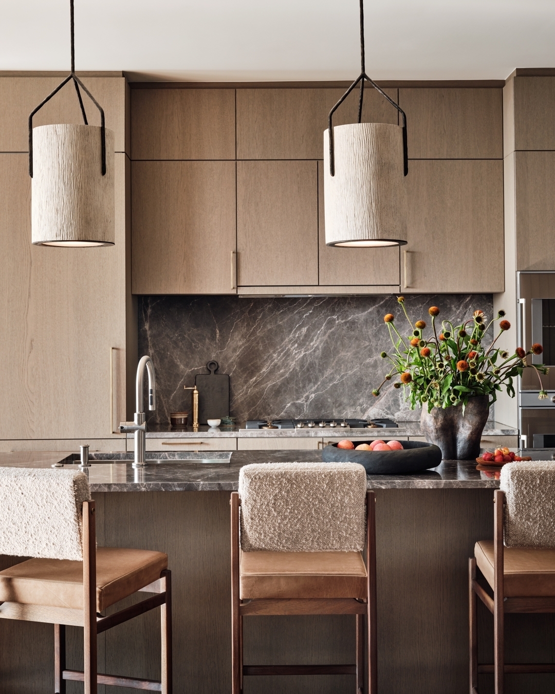 Modern oak kitchen with wooden cabinets, marble backsplash, and countertop. Three upholstered chairs are at the island, with pendant lights above and a vase of flowers and bowl of peaches on the counter—perfect for inspiring kitchen ideas.