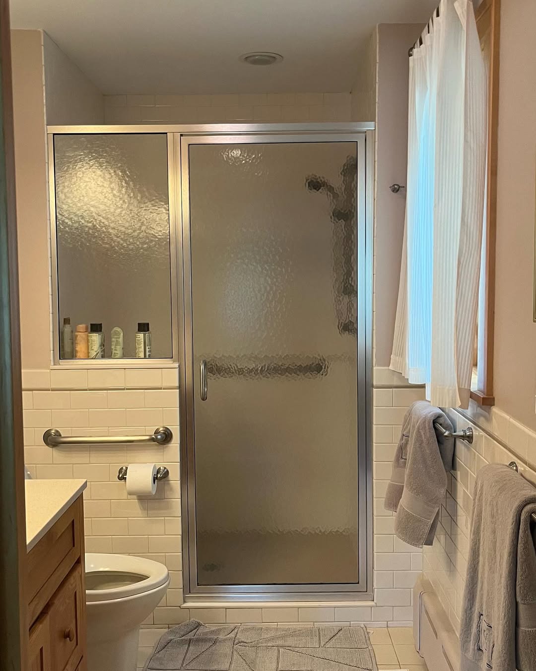 A bathroom featuring a glass shower door with half wall shower ideas, several toiletries on a shelf, a towel rack with towels, a toilet, and a gray bath mat on the tiled floor.
