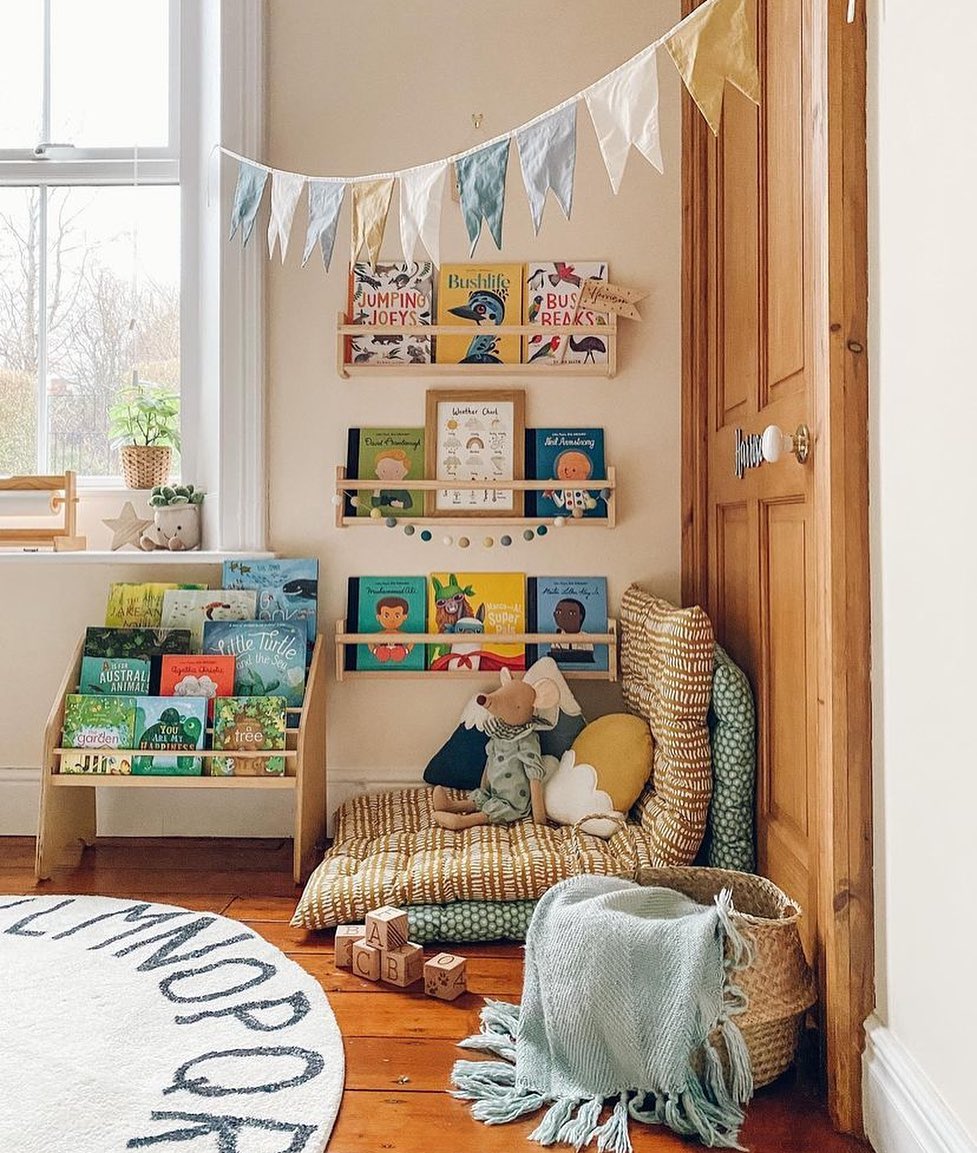 Children’s reading nook with low bookshelves filled with colorful books, floor cushions, a basket with blanket, and decorative bunting above—perfect reading nook inspiration for creating cozy reading corners at home.