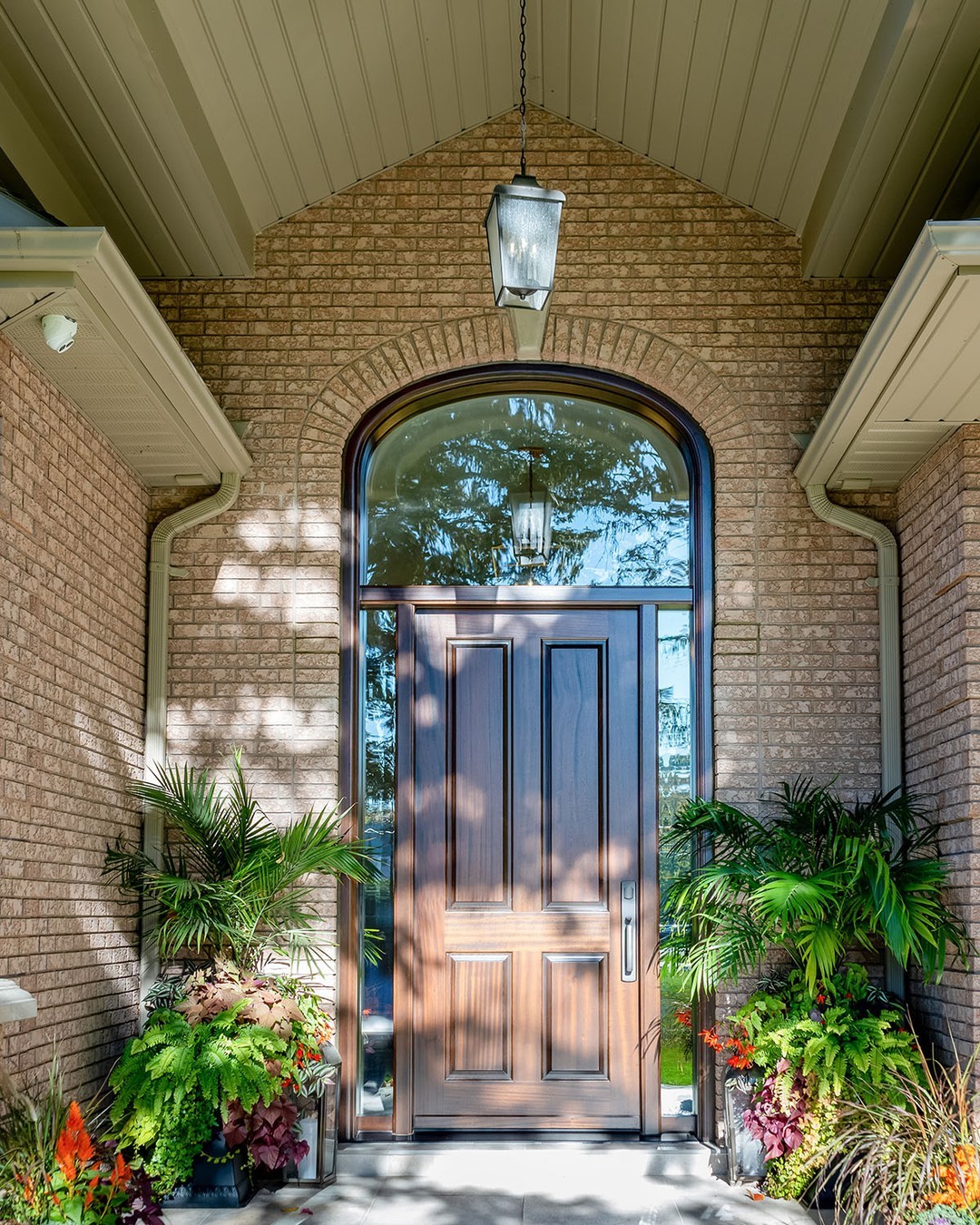 A stylish double door entryway with a large arched window above, flanked by potted plants, set in a brick house exterior under a hanging light fixture—perfect for those seeking elegant entryway ideas.