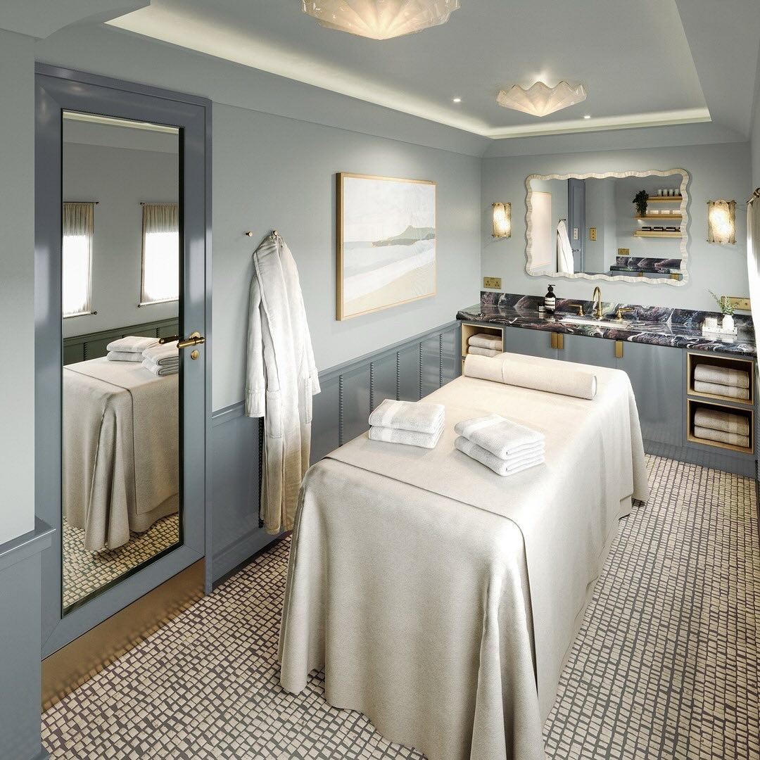A spa treatment room featuring calming neutral tones, thoughtful massage room decor, a massage table, neatly folded towels, a large mirror, wall art, and a stylish sink area.