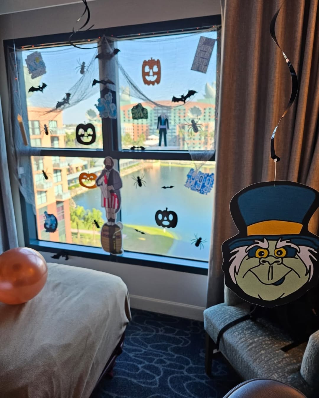 A window decorated with Halloween decorations like pumpkins, bats, and cartoon faces offers great Halloween window decor ideas. Inside are a chair, balloon, and bed, while buildings and water can be seen outside the window.