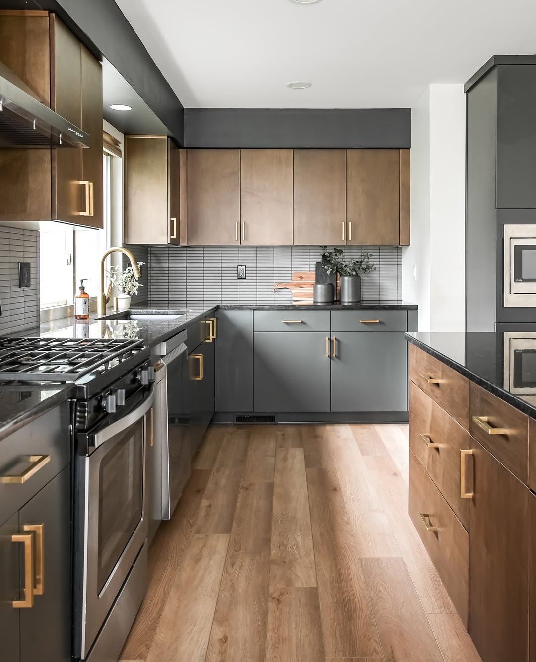 Modern kitchen with stunning kitchen colors, featuring stainless steel appliances, brown and gray cabinets with gold handles, a black countertop, and wood flooring. Small plants and bottles accent the counters.