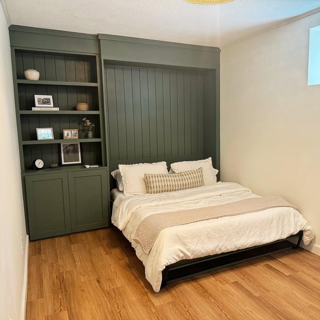 A neatly made Murphy bed with cream bedding is set against a dark green built-in shelving unit with books, photos, and decor, on a wooden floor in a softly lit room.