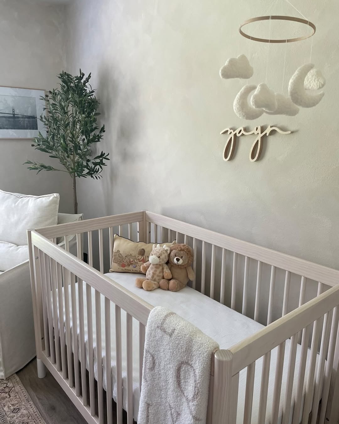 A wooden crib with a white mattress, three stuffed animals, and a blanket sits in a chic nursery. A mobile with clouds and a name hangs above—perfect inspiration for baby boy nursery ideas or any first-time mom.