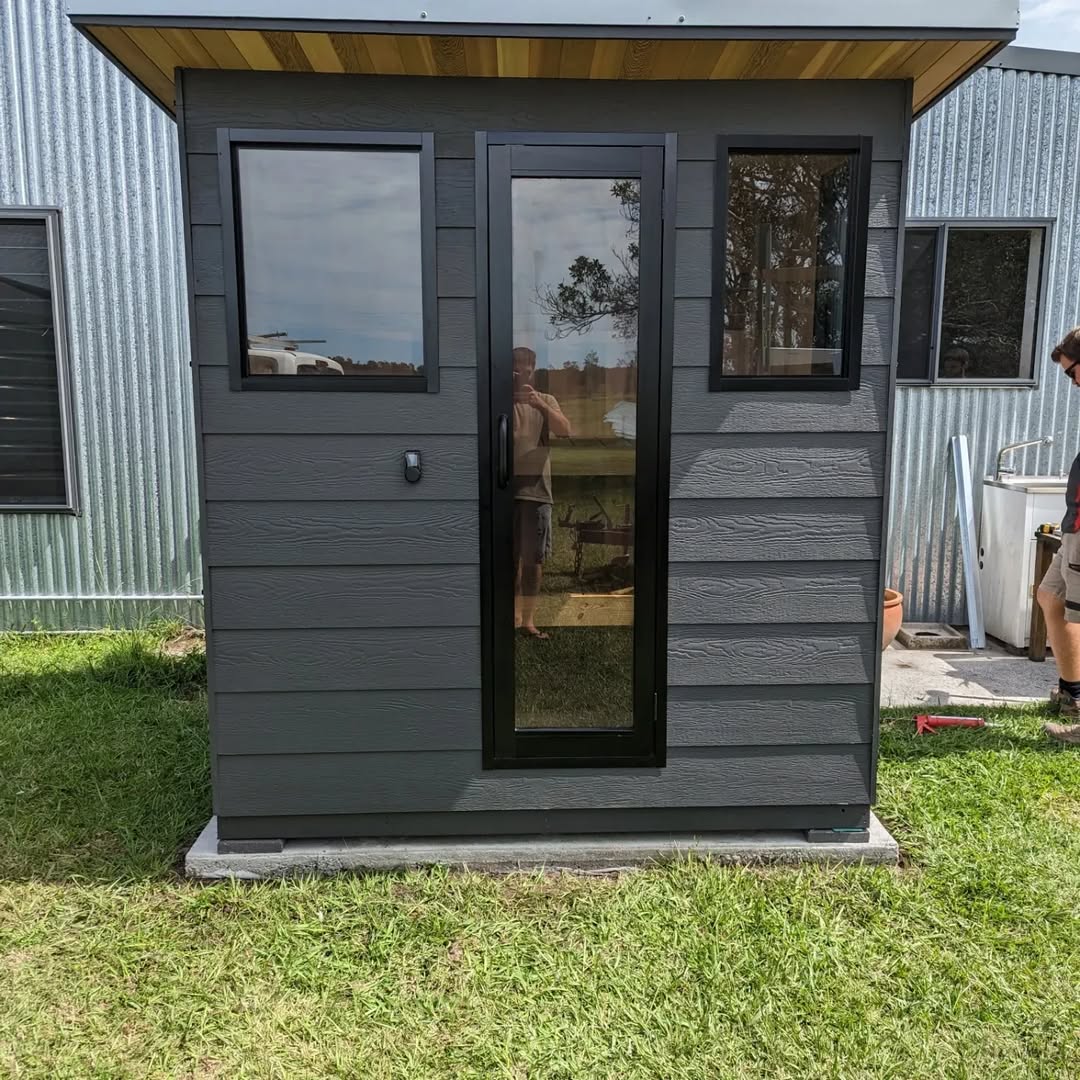 A small, dark gray outdoor sauna with two windows and a glass door stands on a concrete base beside a corrugated metal building on a grassy lawn, offering inspiration for modern sauna design.
