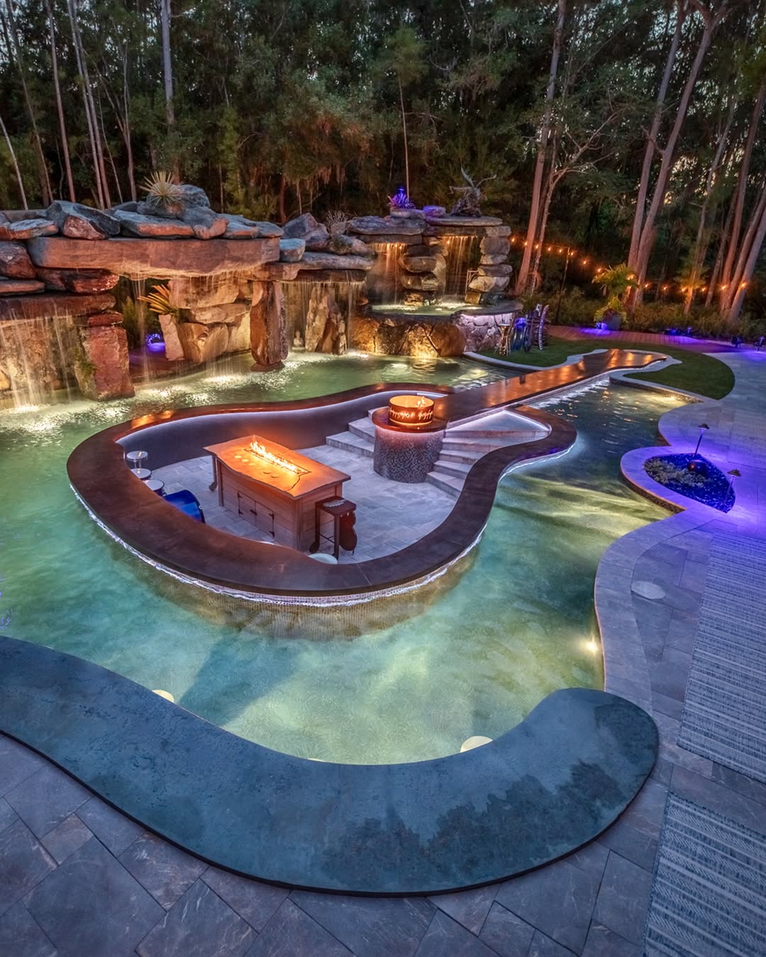 A swimming pool shaped like a guitar features built-in fire pits, a central island with seating, and is surrounded by trees and patio lighting—one of the coolest backyard pool ideas for music lovers.