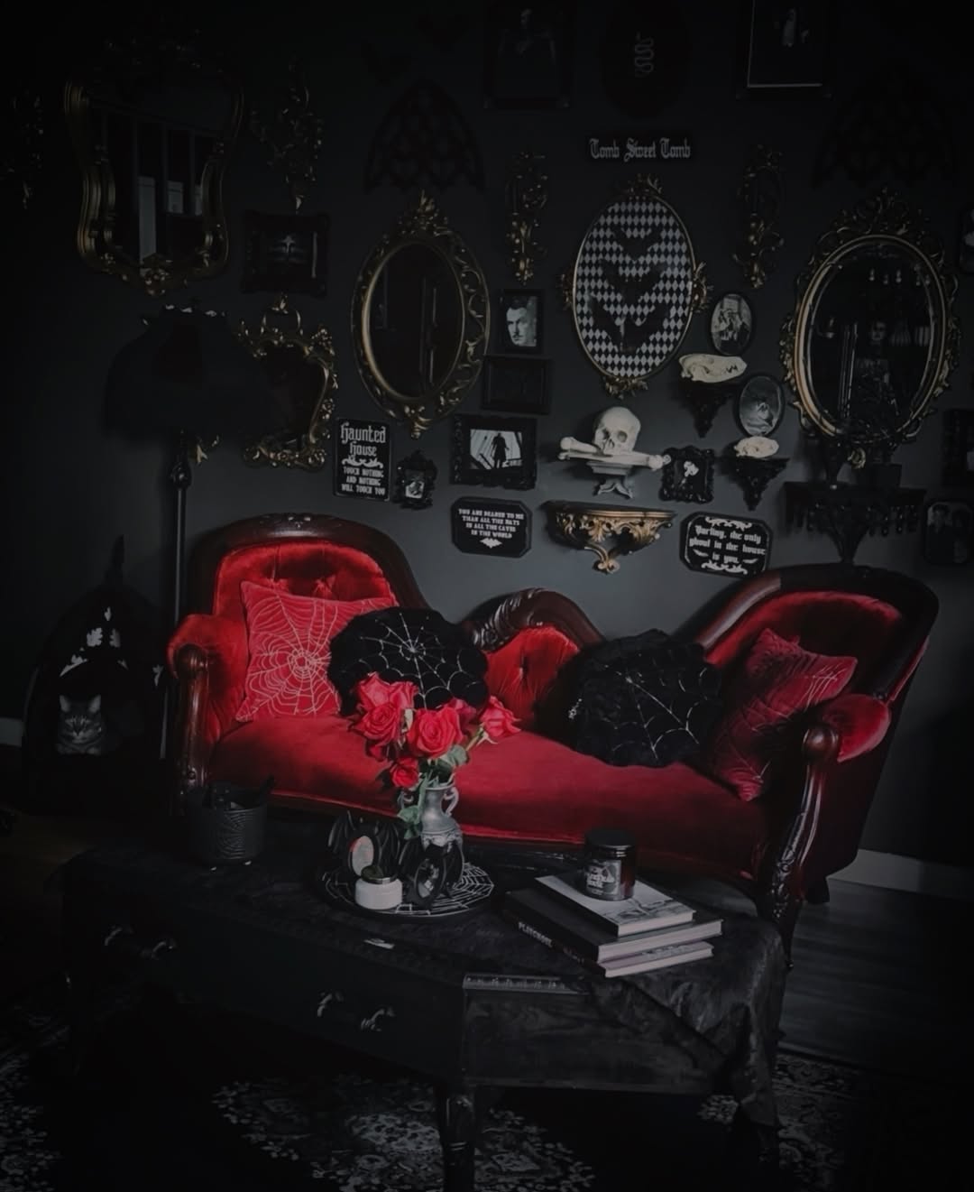 A dark, gothic-themed living room featuring Gothic interior design with a red velvet sofa, black cushions, a coffee table adorned with books and candles, and a wall decorated with mirrors, frames, and macabre art.