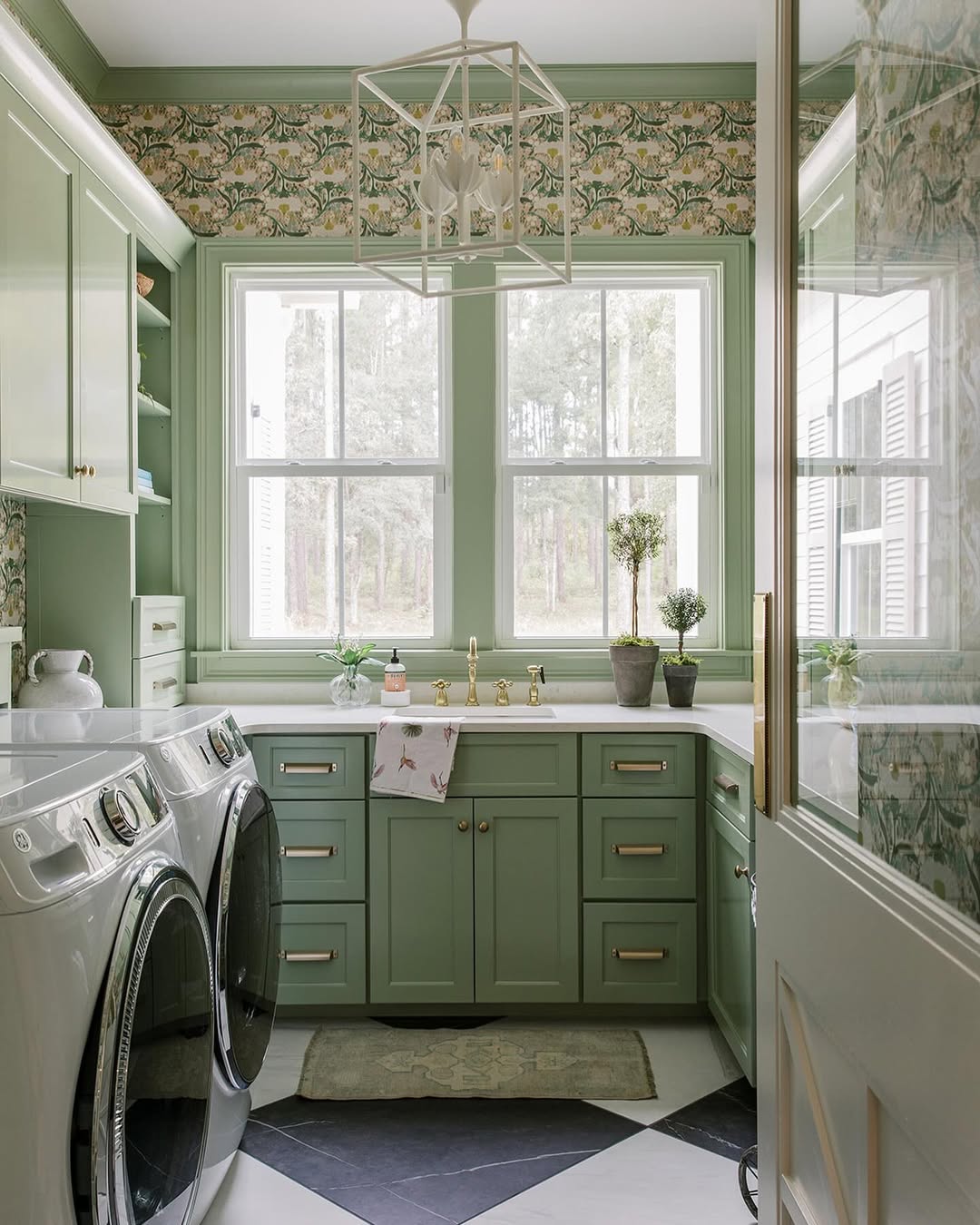 A laundry room with green laundry room cabinets, a washer and dryer, a countertop with potted plants, and large windows letting in natural light—perfect for those seeking fresh cabinet ideas.