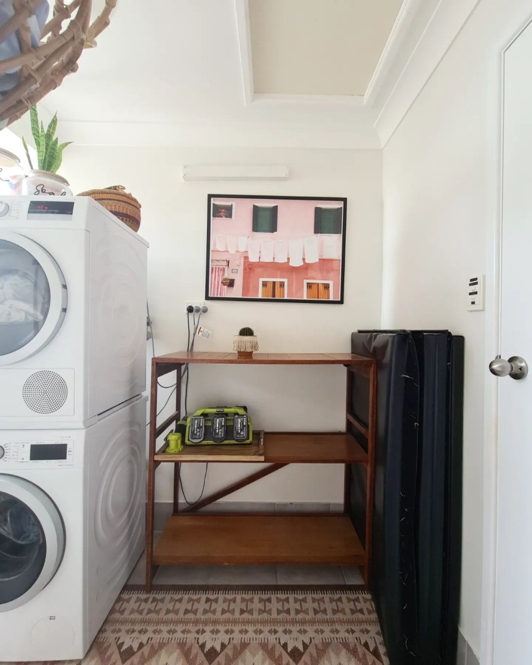 A laundry room with a stacked washer and dryer, a wooden shelf holding battery chargers, a folded mattress, and a framed photo of a pink building with laundry hanging—perfect for those seeking boho laundry room inspiration.