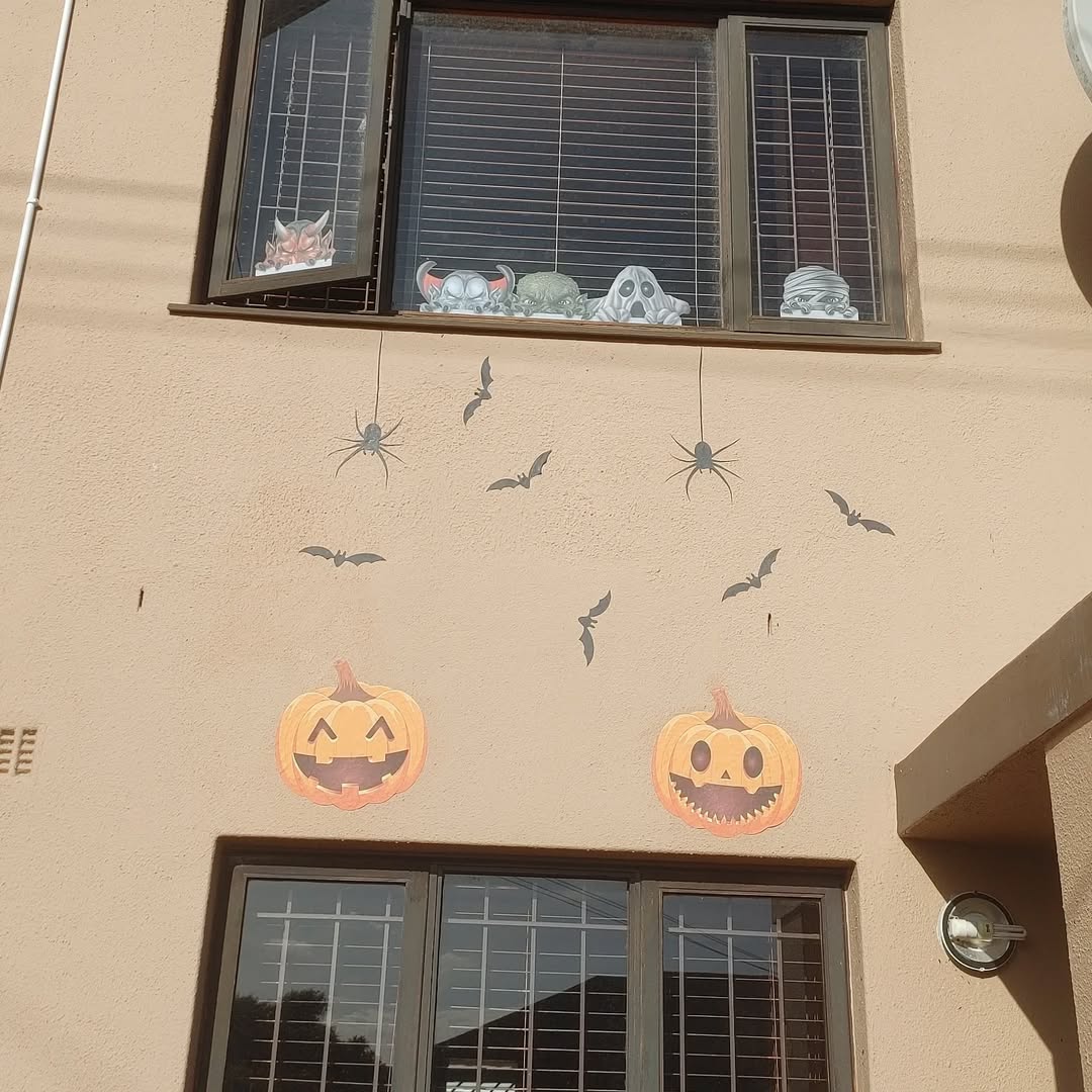 Two windows on a beige wall feature creative Halloween window decoration ideas with masks, paper bats, spiders, and two large paper jack-o’-lanterns.